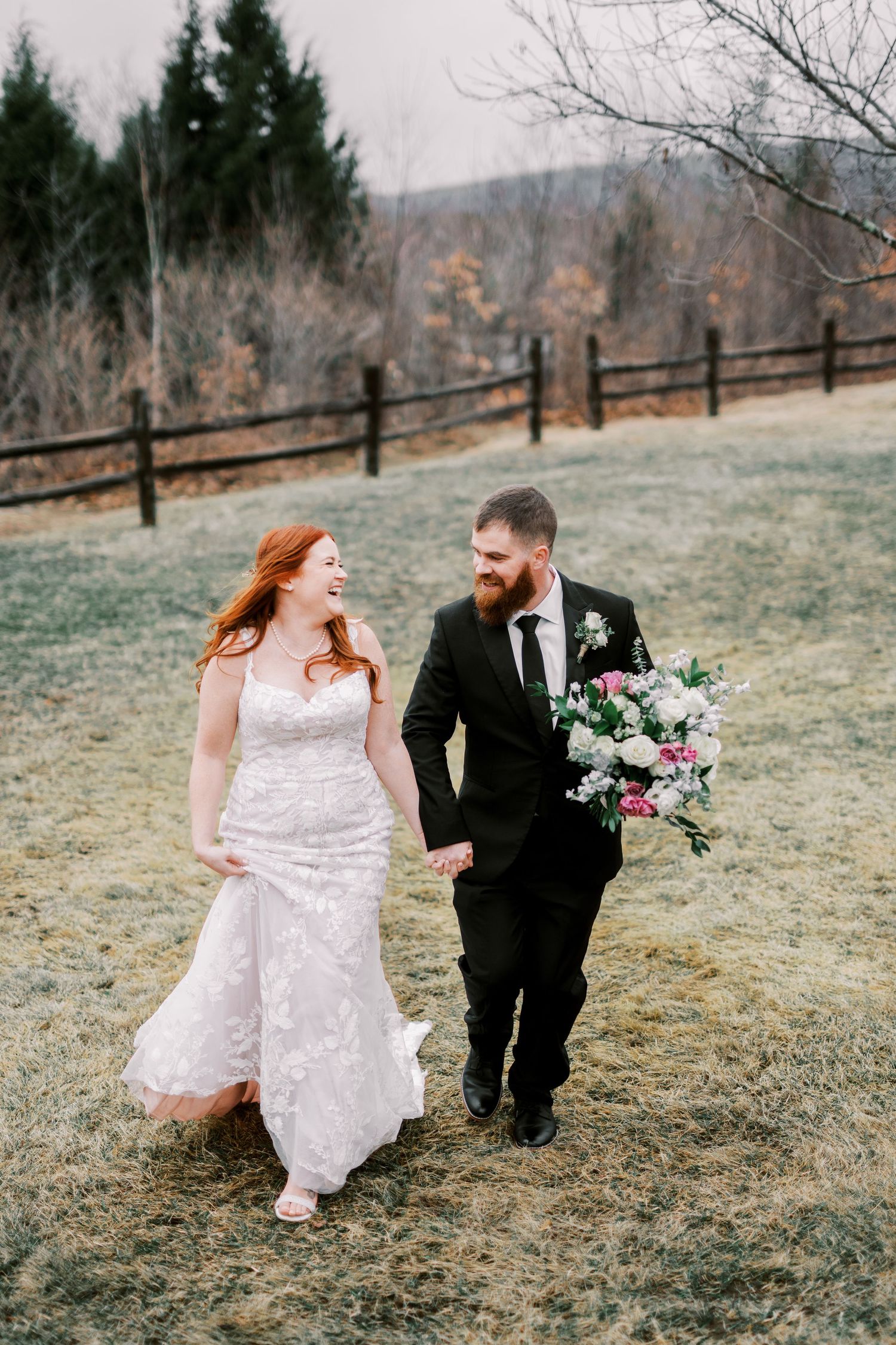 A couple shares a joyful moment walking together on a hillside with winter trees and fencing in the background.