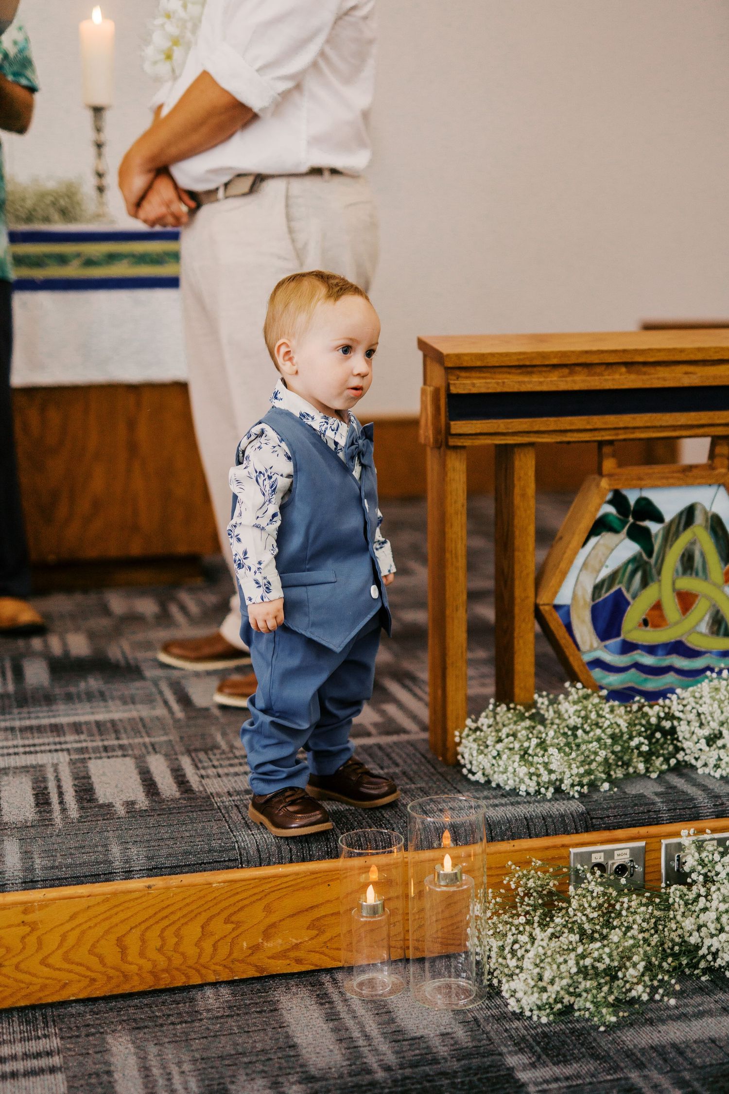 Small child in blue vest standing on wooden platform near floral decorations.