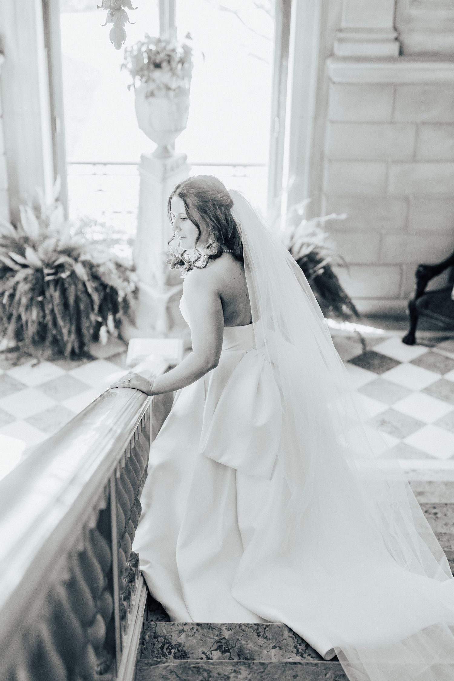 Elegant bride in white wedding gown and veil standing by window in classic indoor setting.