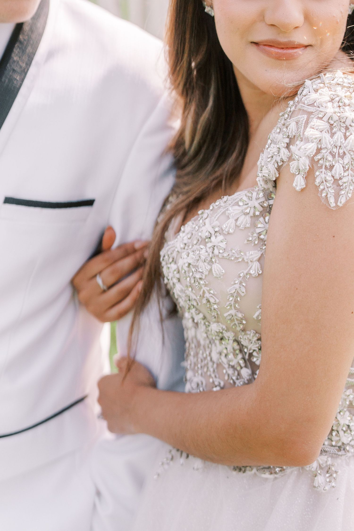 Close-up detail of a bride wearing an ornate beaded wedding gown standing next to someone in a white suit jacket.