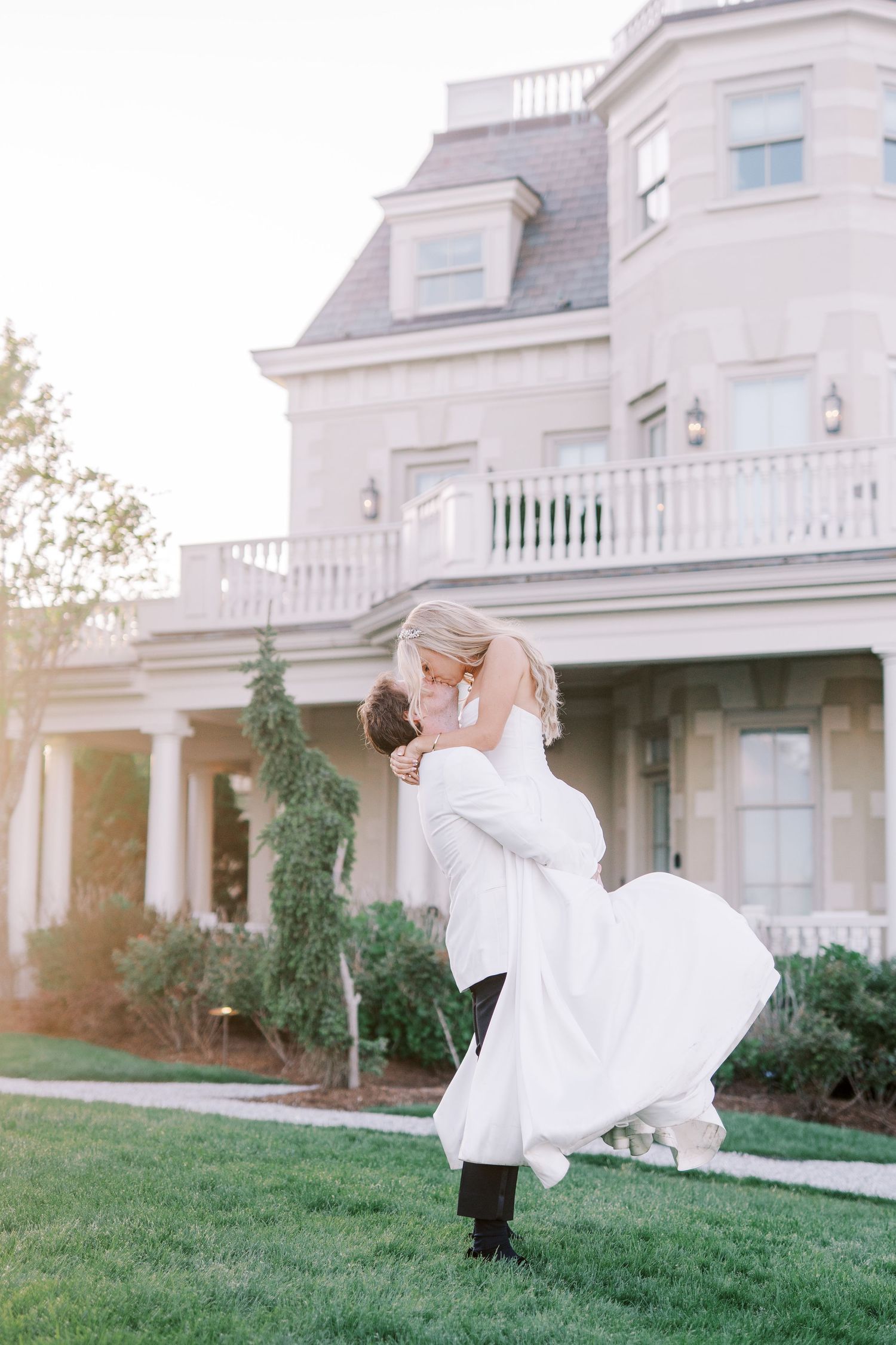 A romantic wedding scene outside a stately white mansion with lush landscaping and elegant architectural details.