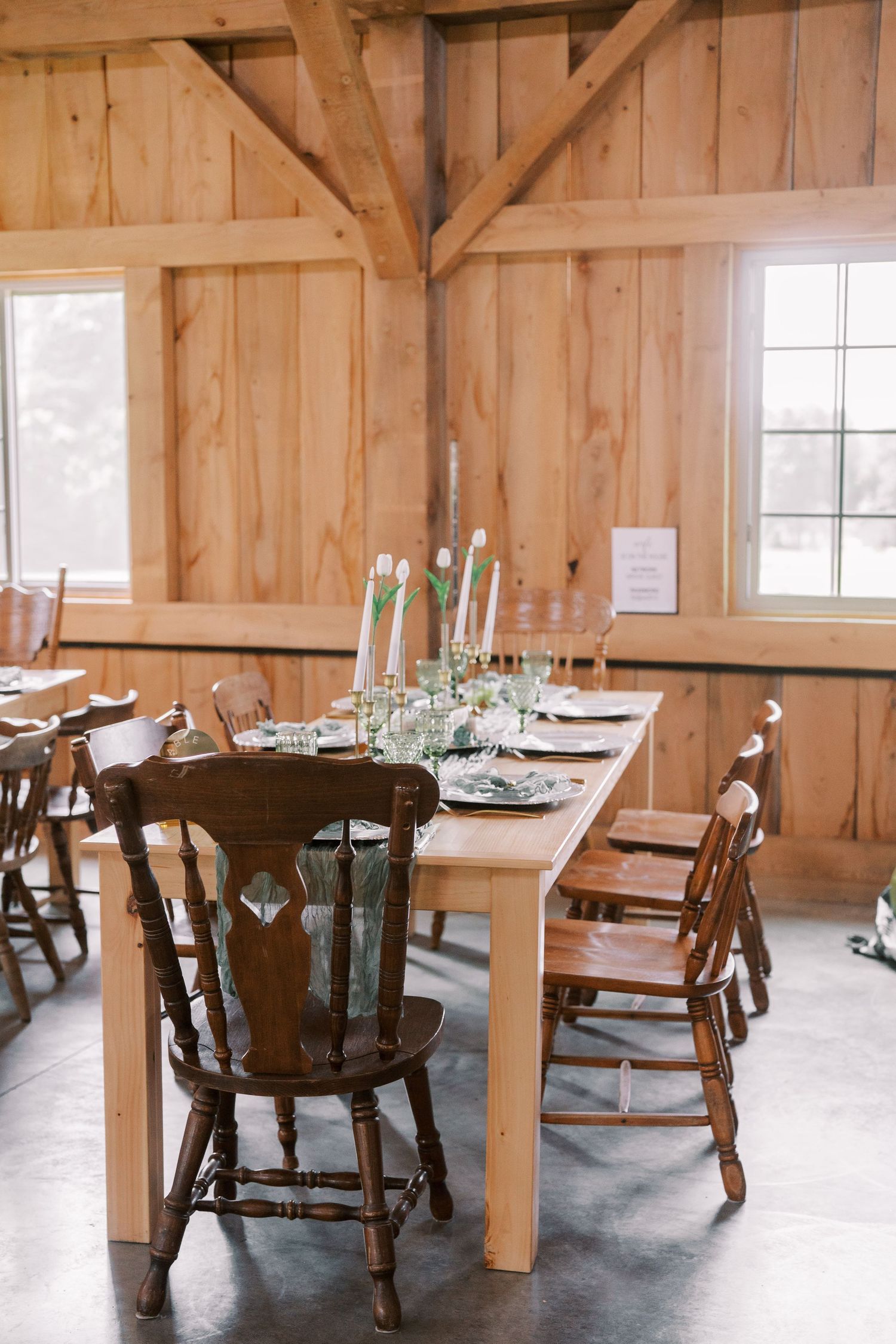 Rustic wooden dining table set with antique chairs in a barn-style room with natural wood walls and white candles.