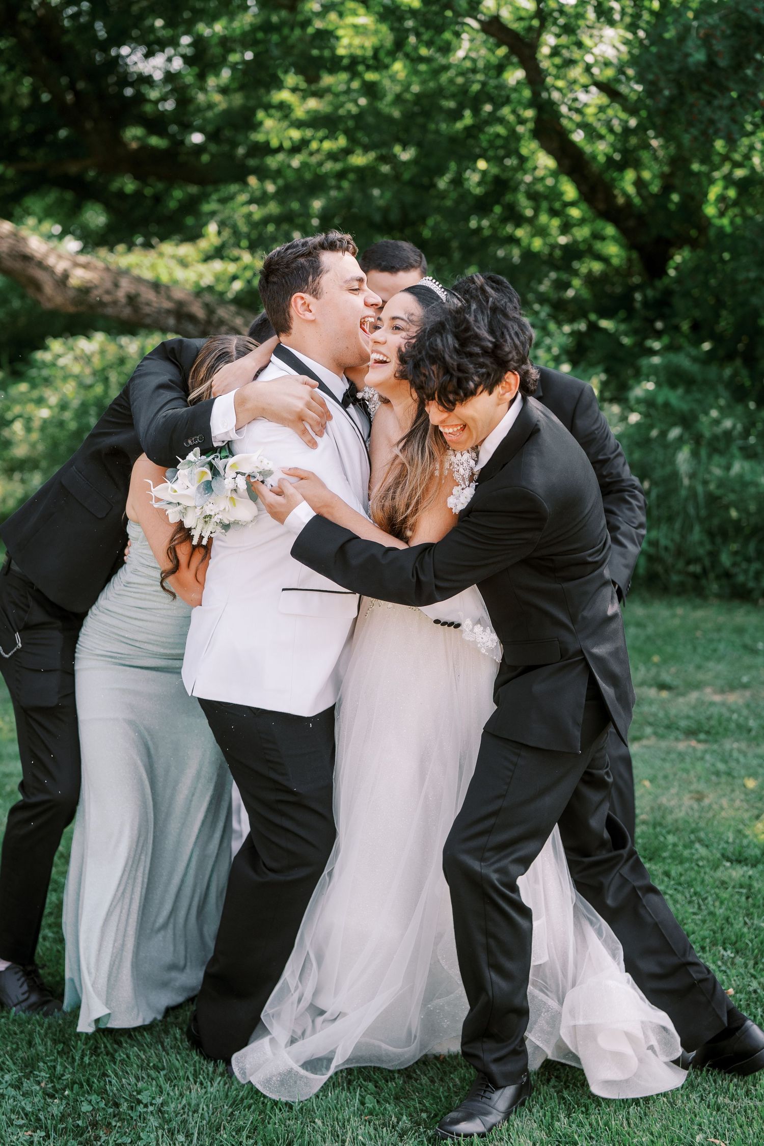 A joyful group hug among wedding party members on a grassy lawn with green trees in the background.