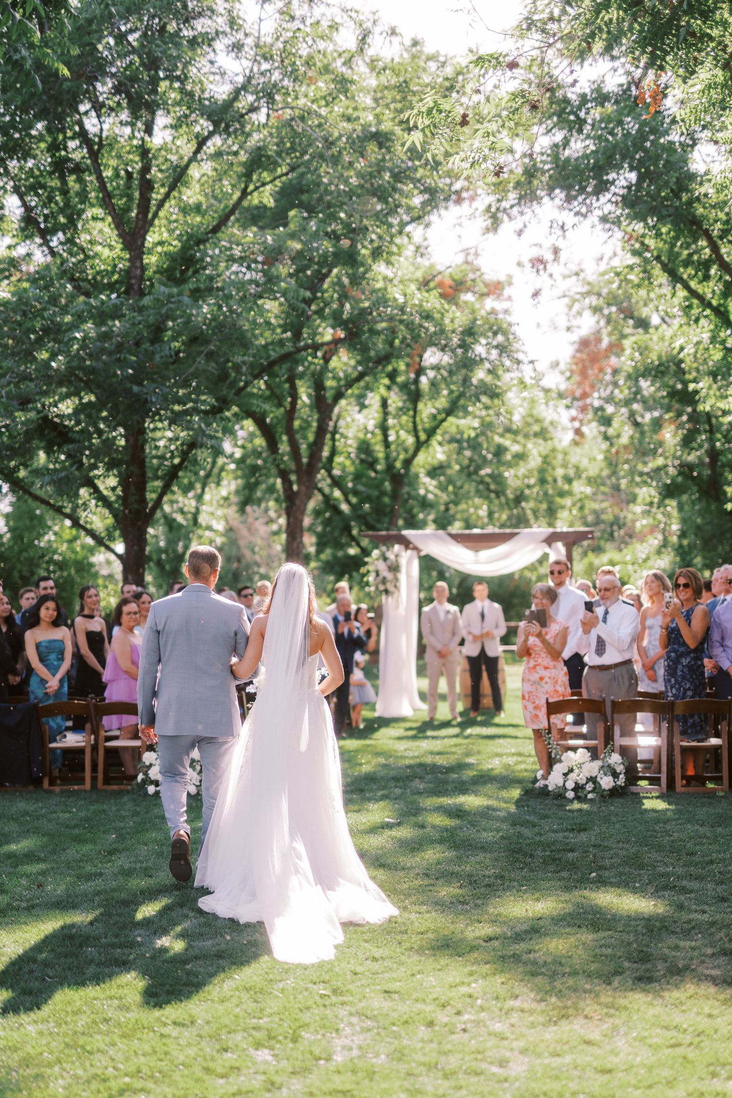 A beautiful outdoor garden wedding ceremony under tall trees with guests seated on wooden chairs and a white fabric arch.
