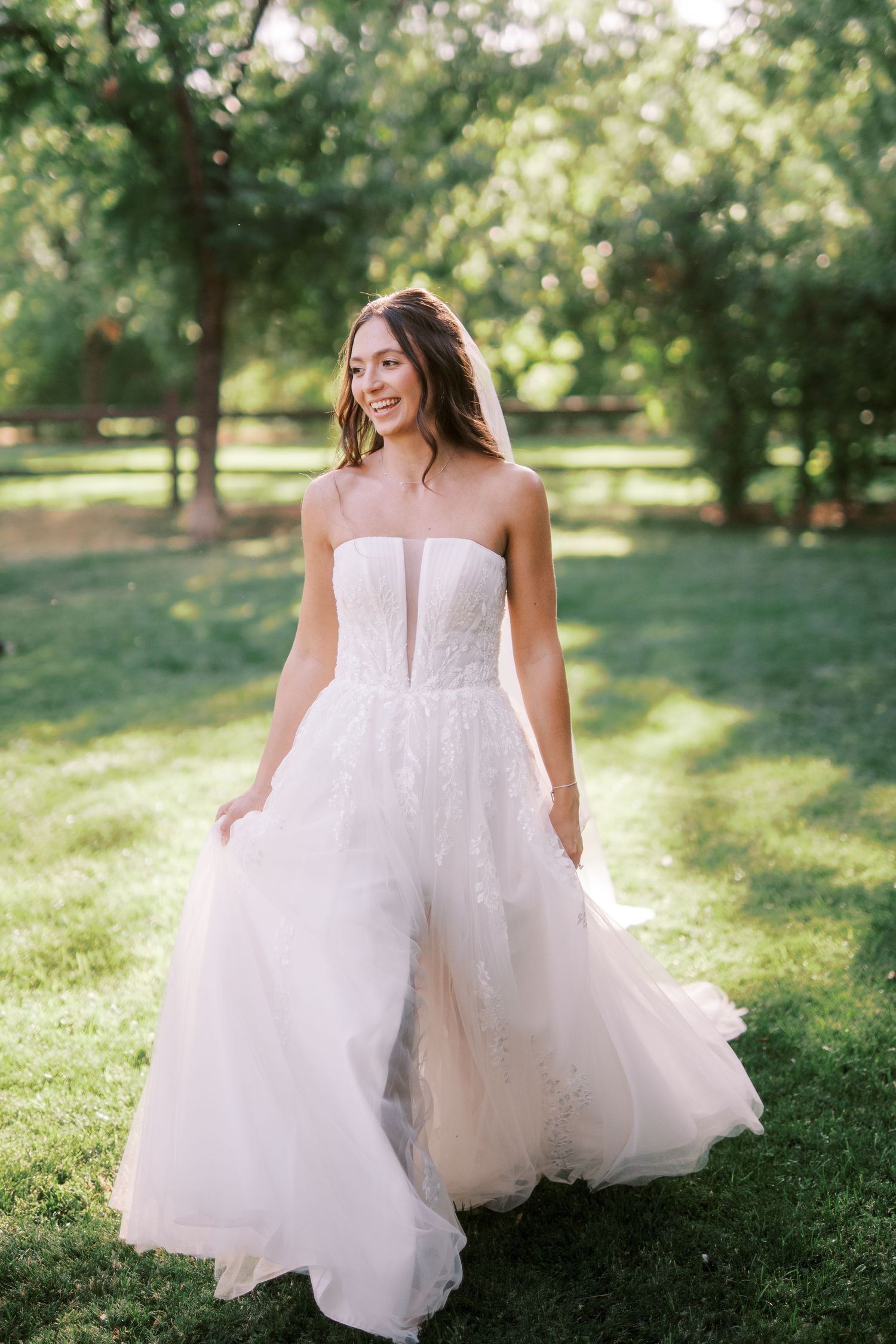 A woman in a flowing white wedding dress spins joyfully on green grass in a sunlit garden setting.
