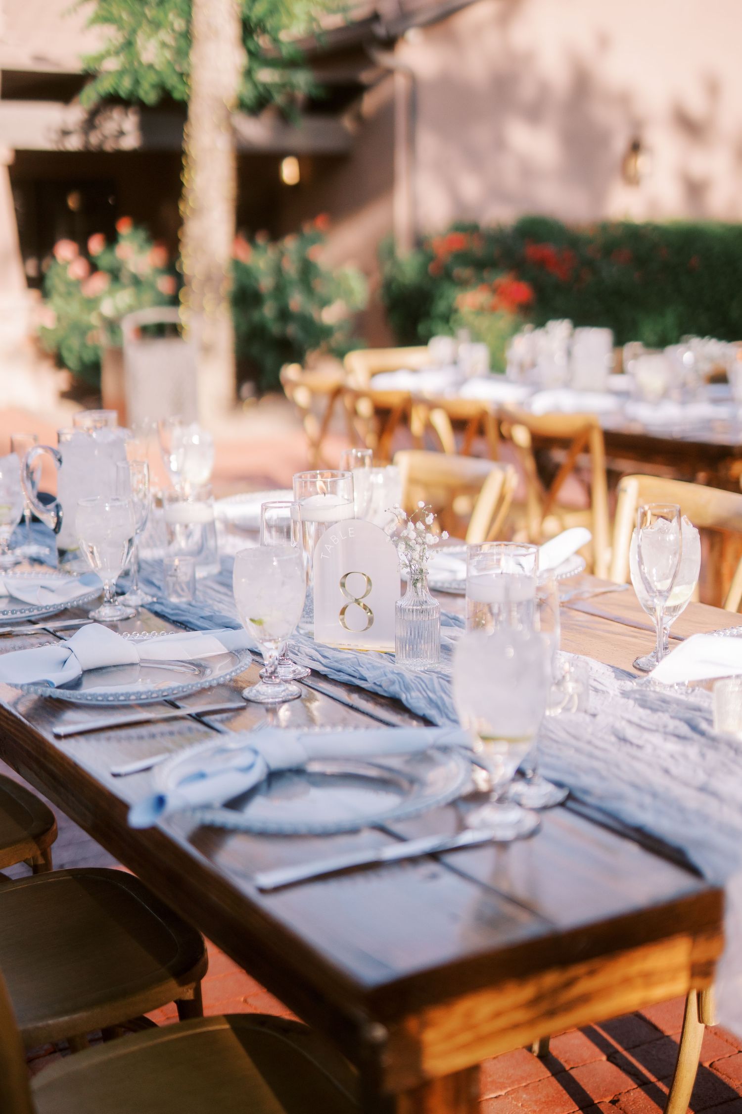 Elegant outdoor wedding reception tables set with light blue linens, charger plates, and glassware in a garden setting.