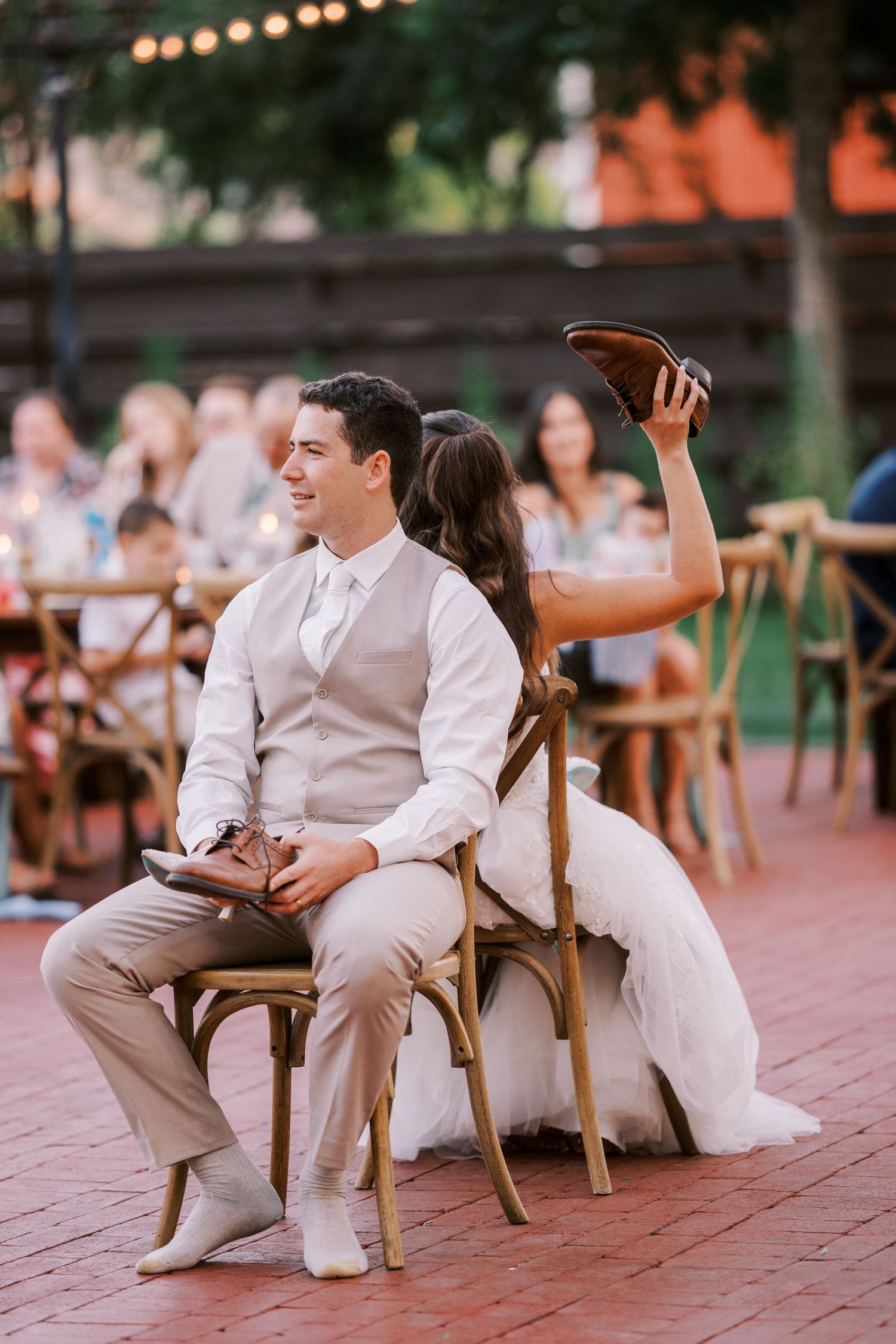 Couple plays wedding shoe game on rustic wooden chairs at outdoor reception under string lights.