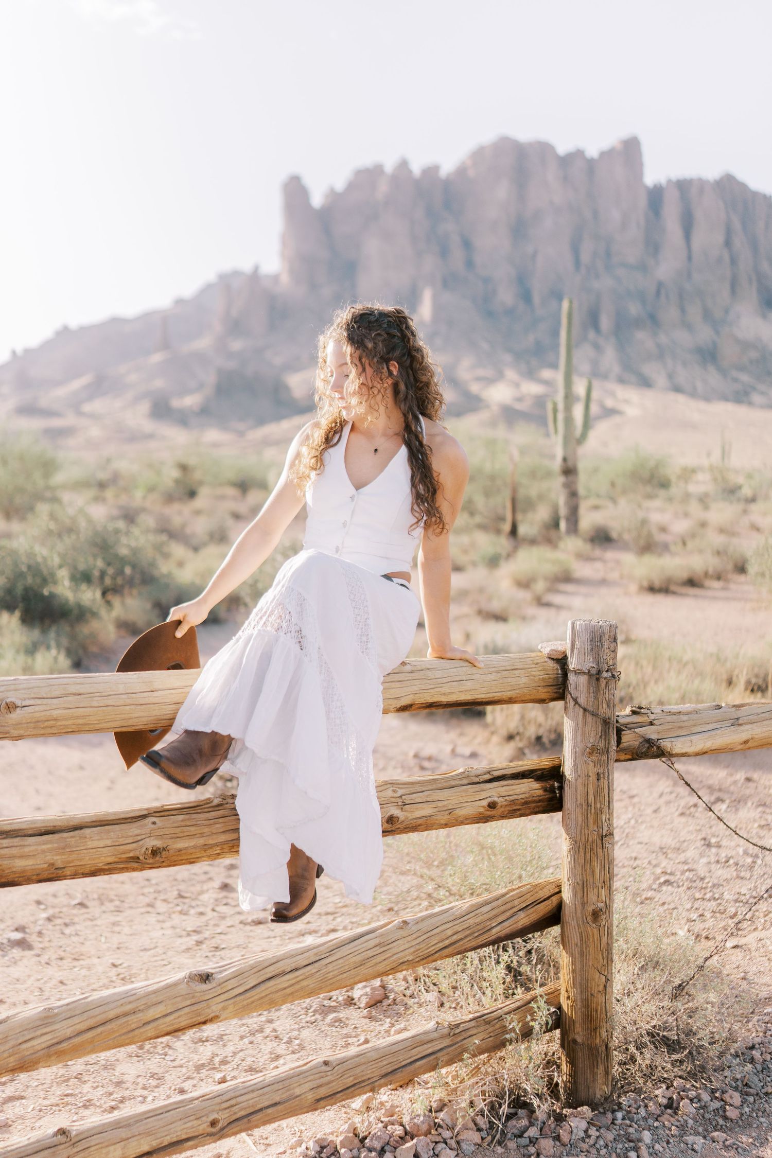 A woman in a flowing white dress sits on a wooden fence in the Arizona desert with mountains and saguaro cactus in background.