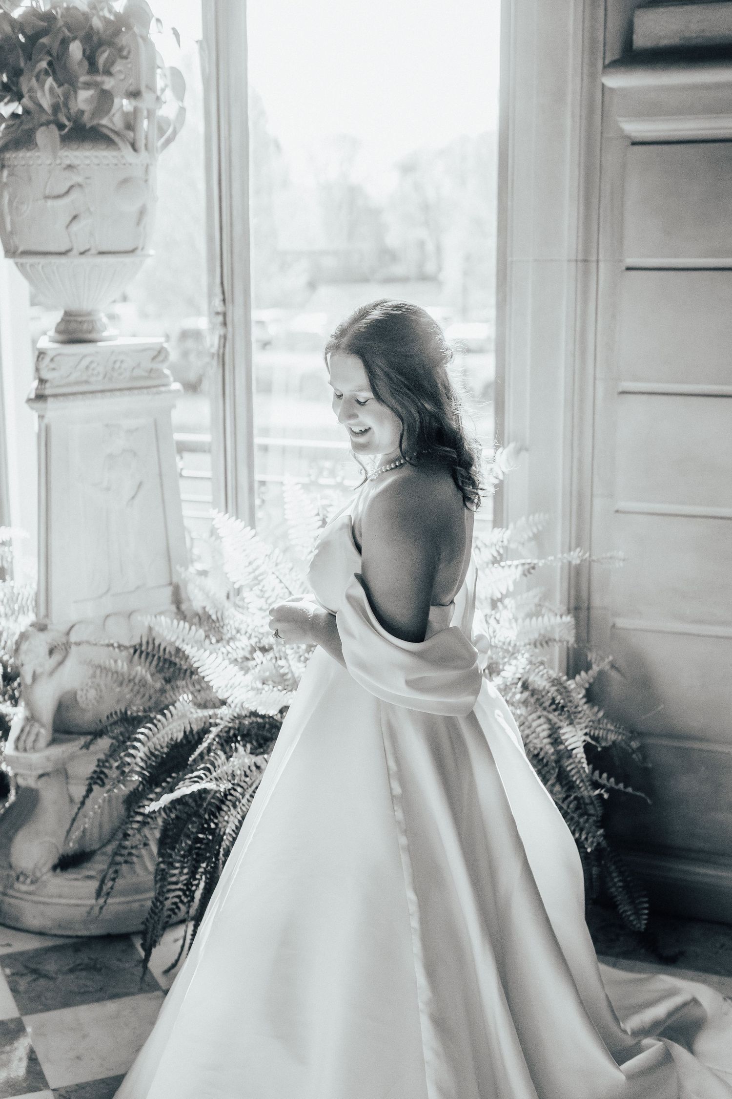 Black and white photograph of a bride in an elegant satin ball gown standing by a window with ferns and natural light.