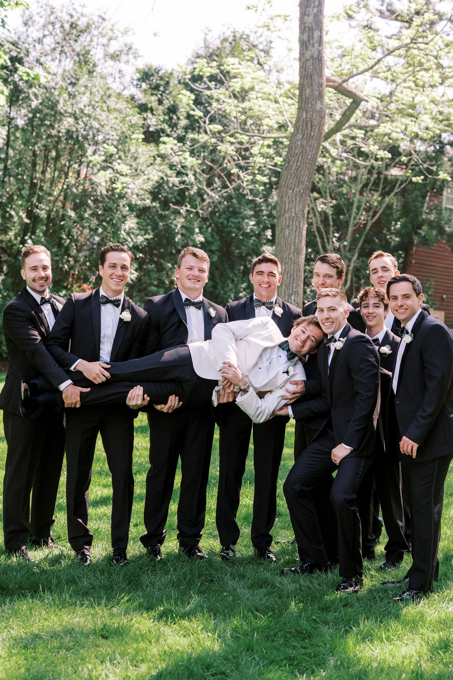 A wedding party in black tuxedos poses playfully on a grassy lawn while lifting one member of the group.