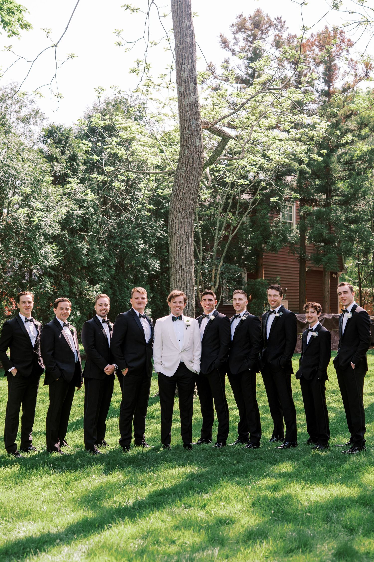 Wedding party in formal attire poses on green lawn with trees in background, groom stands out in white jacket.