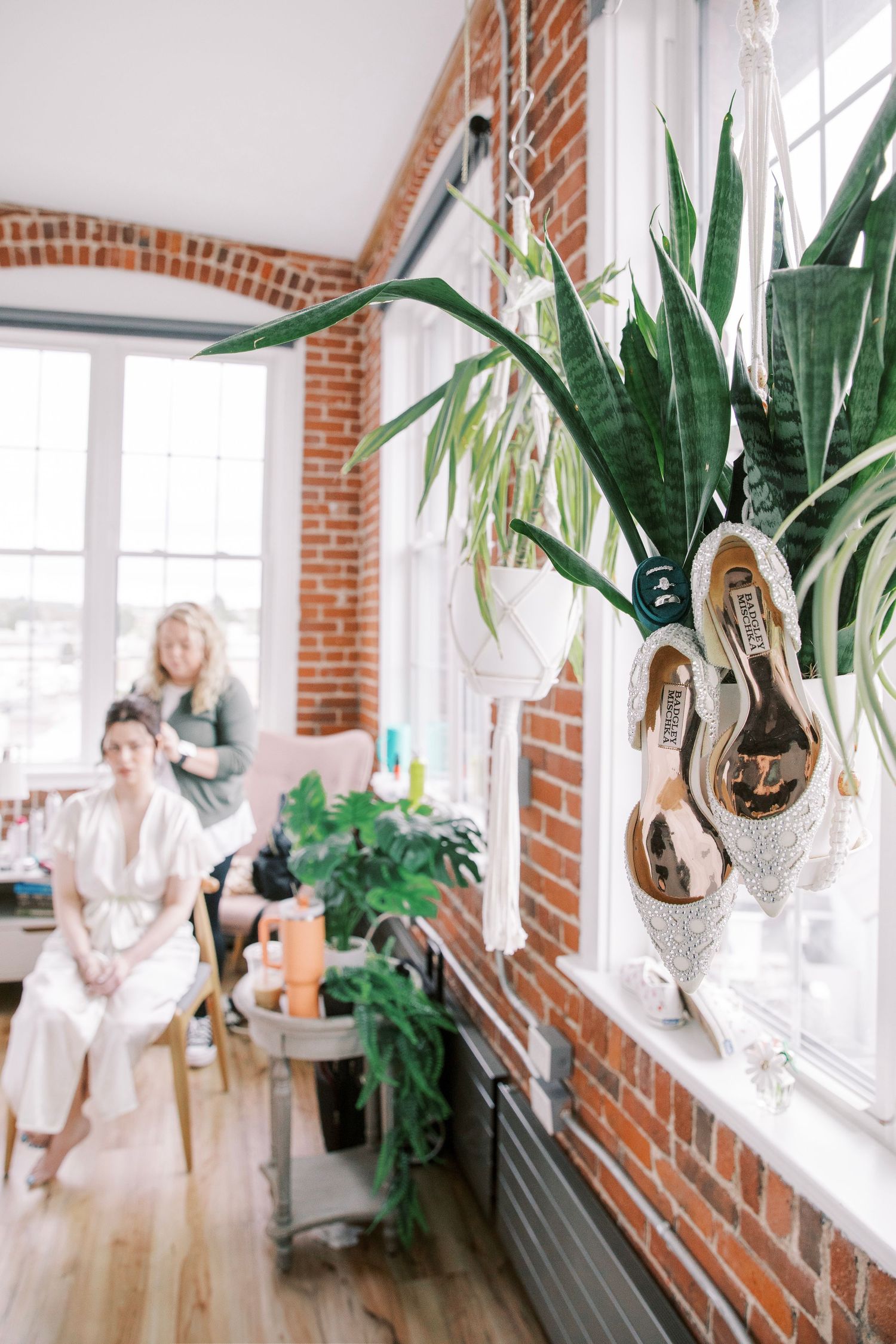 Decorative wedding shoes hang in window with plants and exposed brick interior of bridal suite.