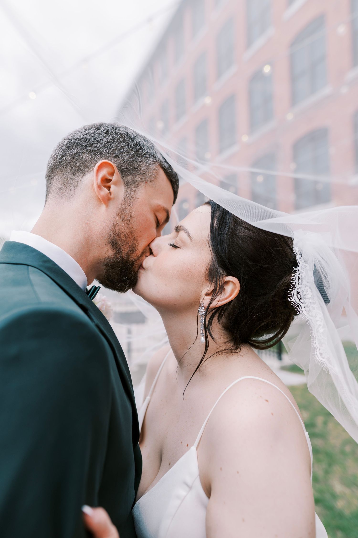 Intimate wedding portrait with flowing veil against urban brick building backdrop.