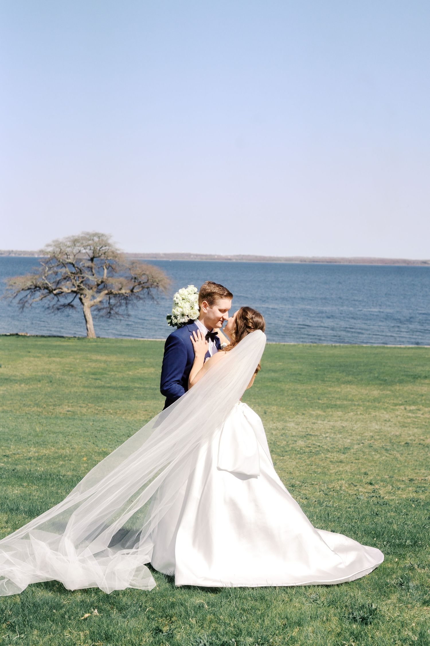 A wedding couple embraces on a scenic lakefront lawn with a lone tree in the background.