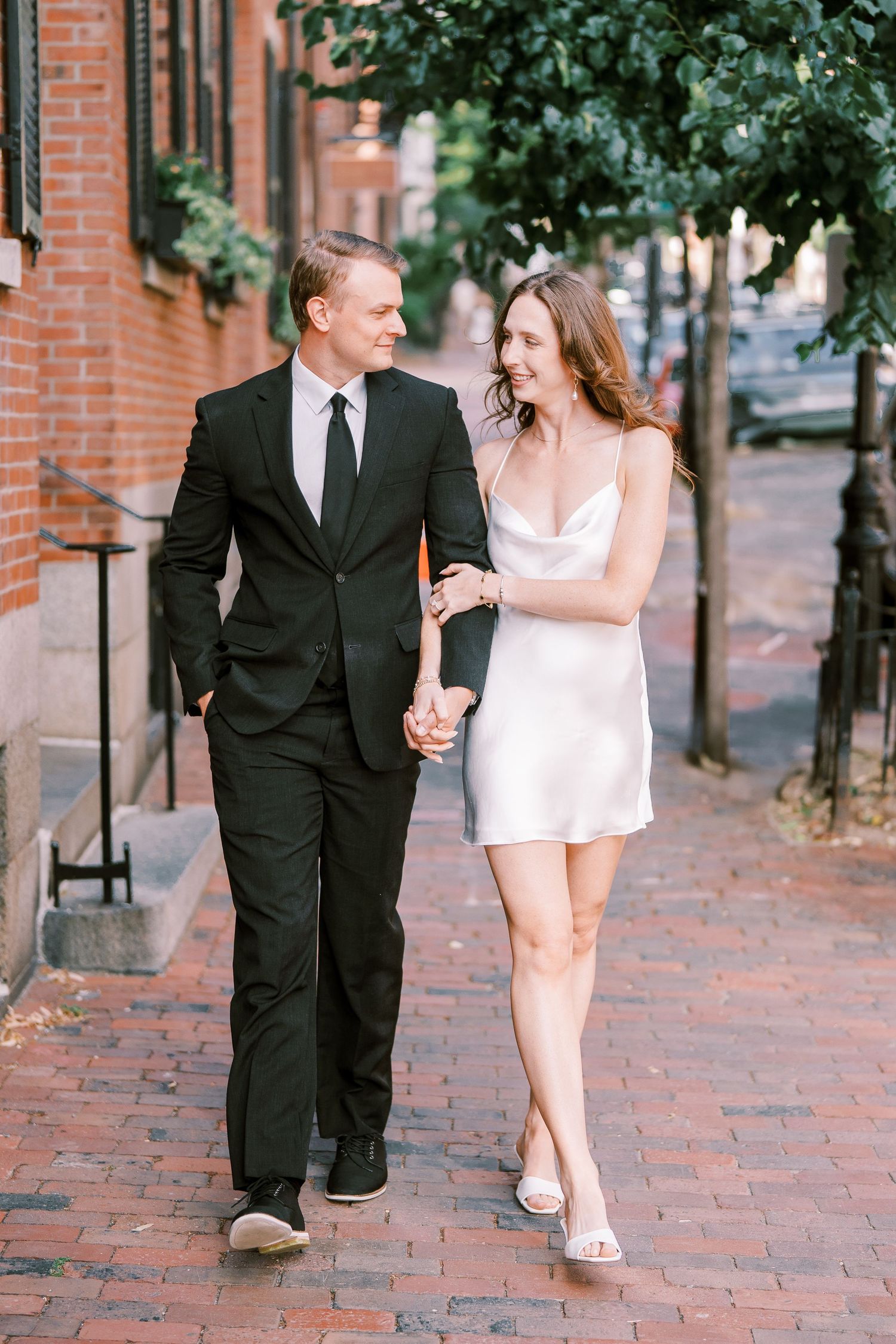 Couple in formal attire walks and laughs together on a brick sidewalk in front of historic buildings in three similar poses.