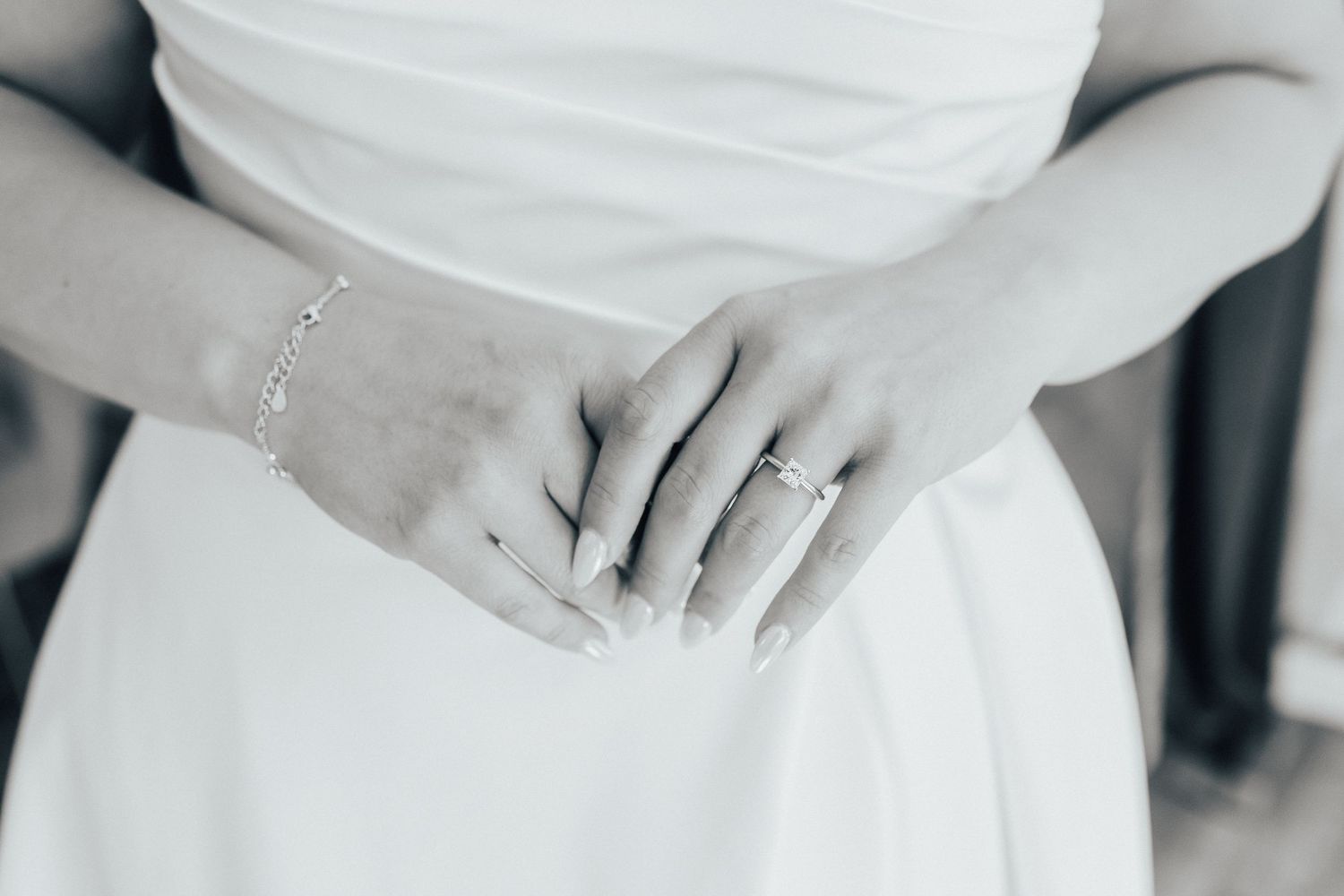 Close-up black and white photo of hands wearing delicate jewelry against a white dress.