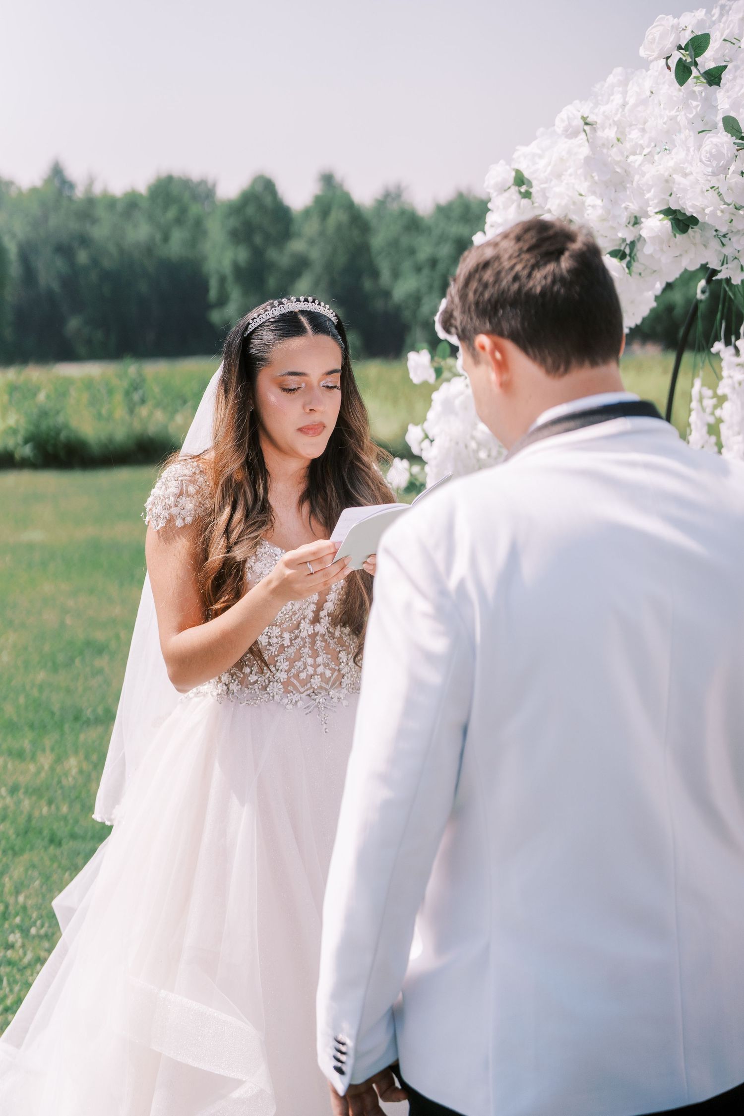 A couple exchanges vows during outdoor wedding ceremony with decorative white floral arrangements and forest backdrop.