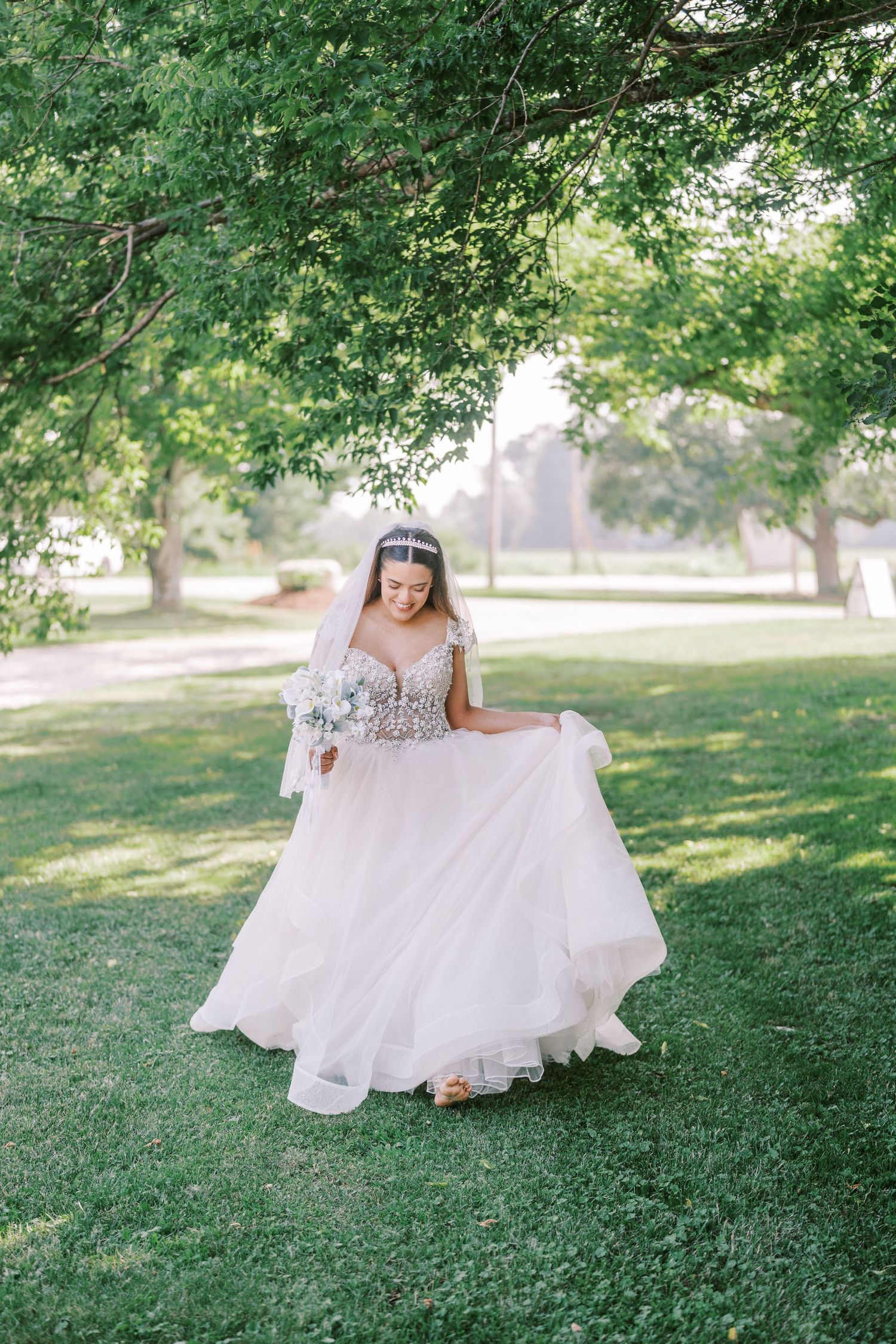 Elegant bride in flowing white wedding dress spins gracefully on green lawn under sunlit tree branches.