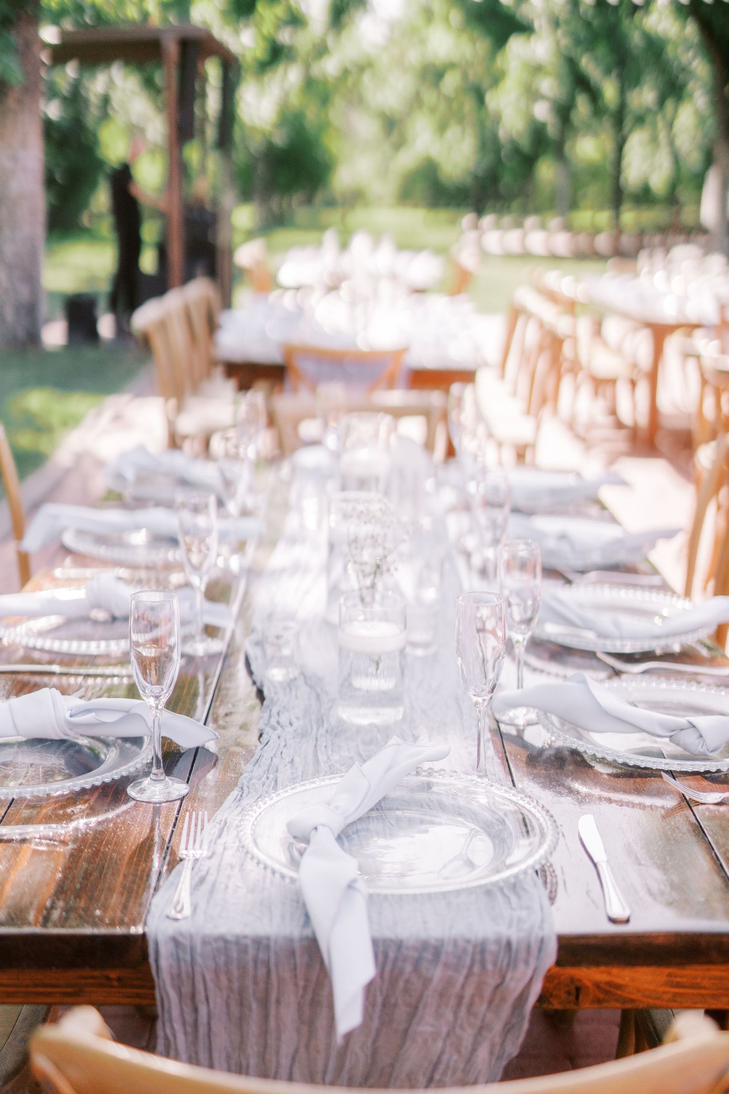 Elegant outdoor wedding reception table setting with white linens and wooden chairs in a garden setting.