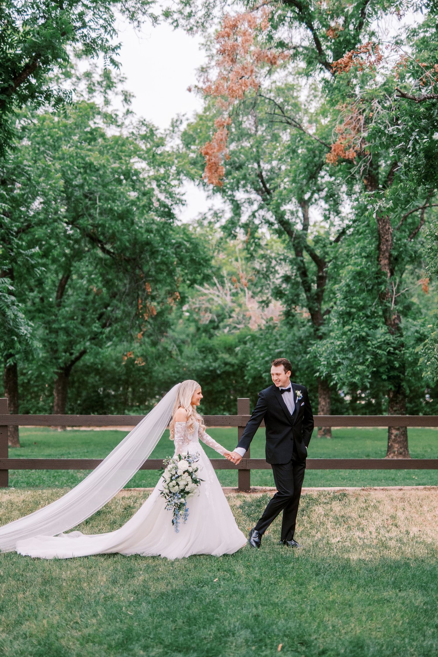 A couple in wedding attire strolls hand in hand through a lush garden setting with flowing veil and green trees.
