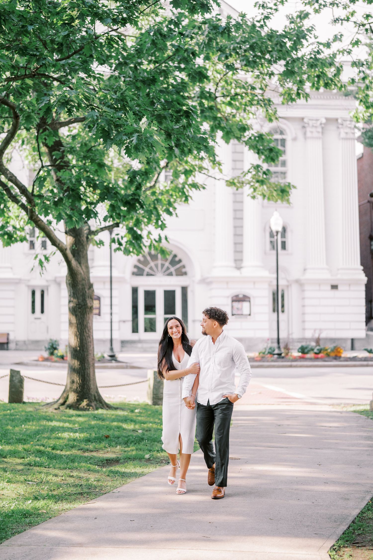 A couple walks together on a path near a grand white classical building on a sunny day.