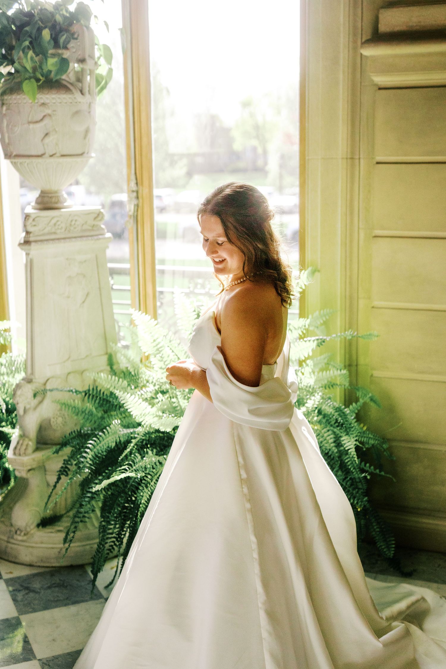 Elegant white wedding gown with off-shoulder design photographed in a sunlit room with decorative ferns and classical architecture.