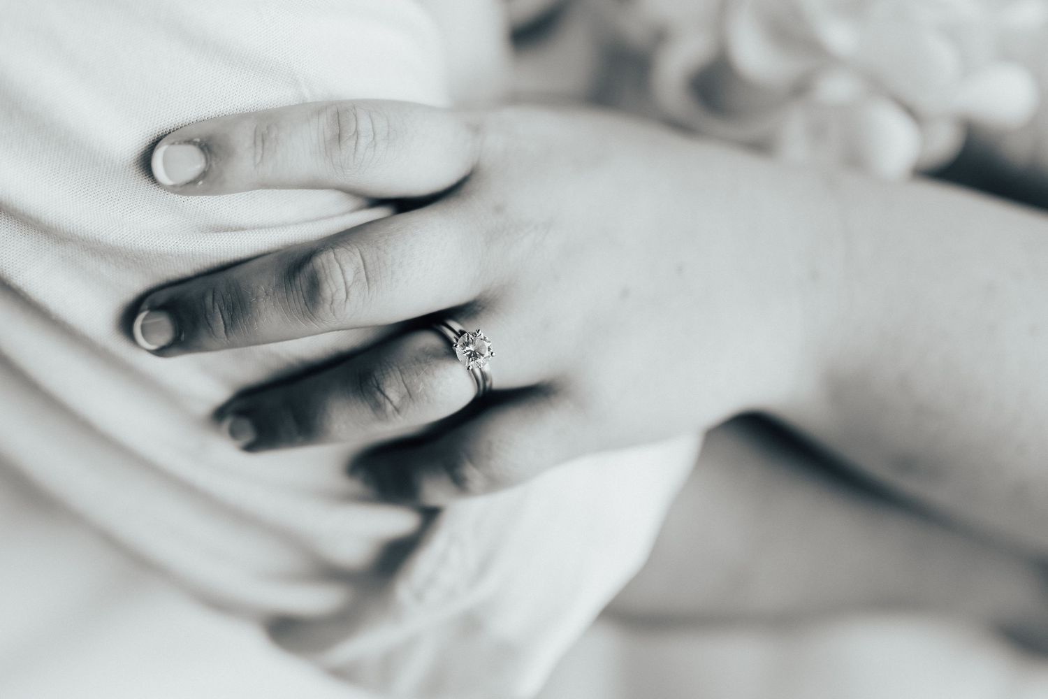Close-up shot of a hand wearing an engagement ring against a light fabric background.