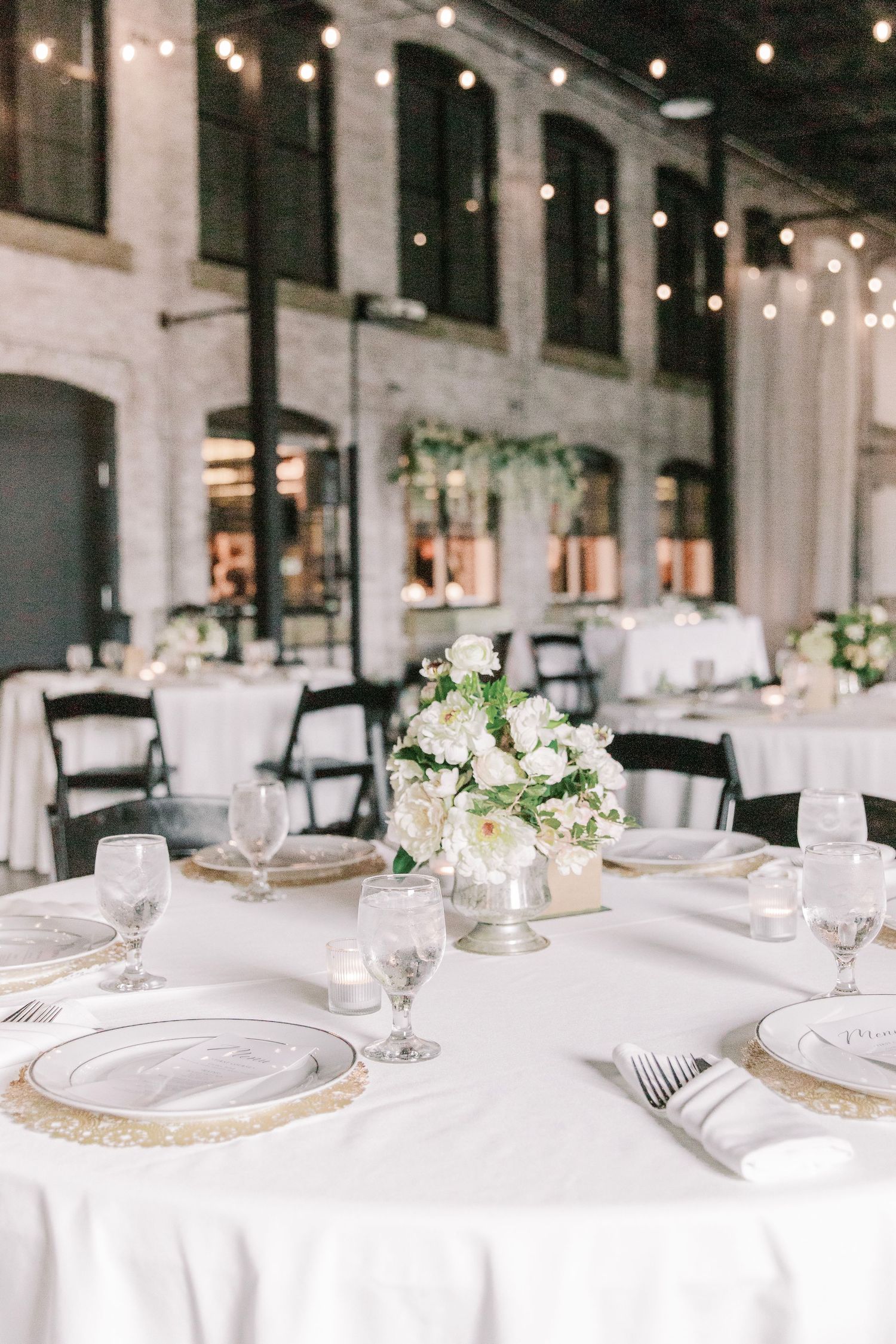 Elegant wedding reception setup with white linens and floral centerpieces in an industrial-chic venue.
