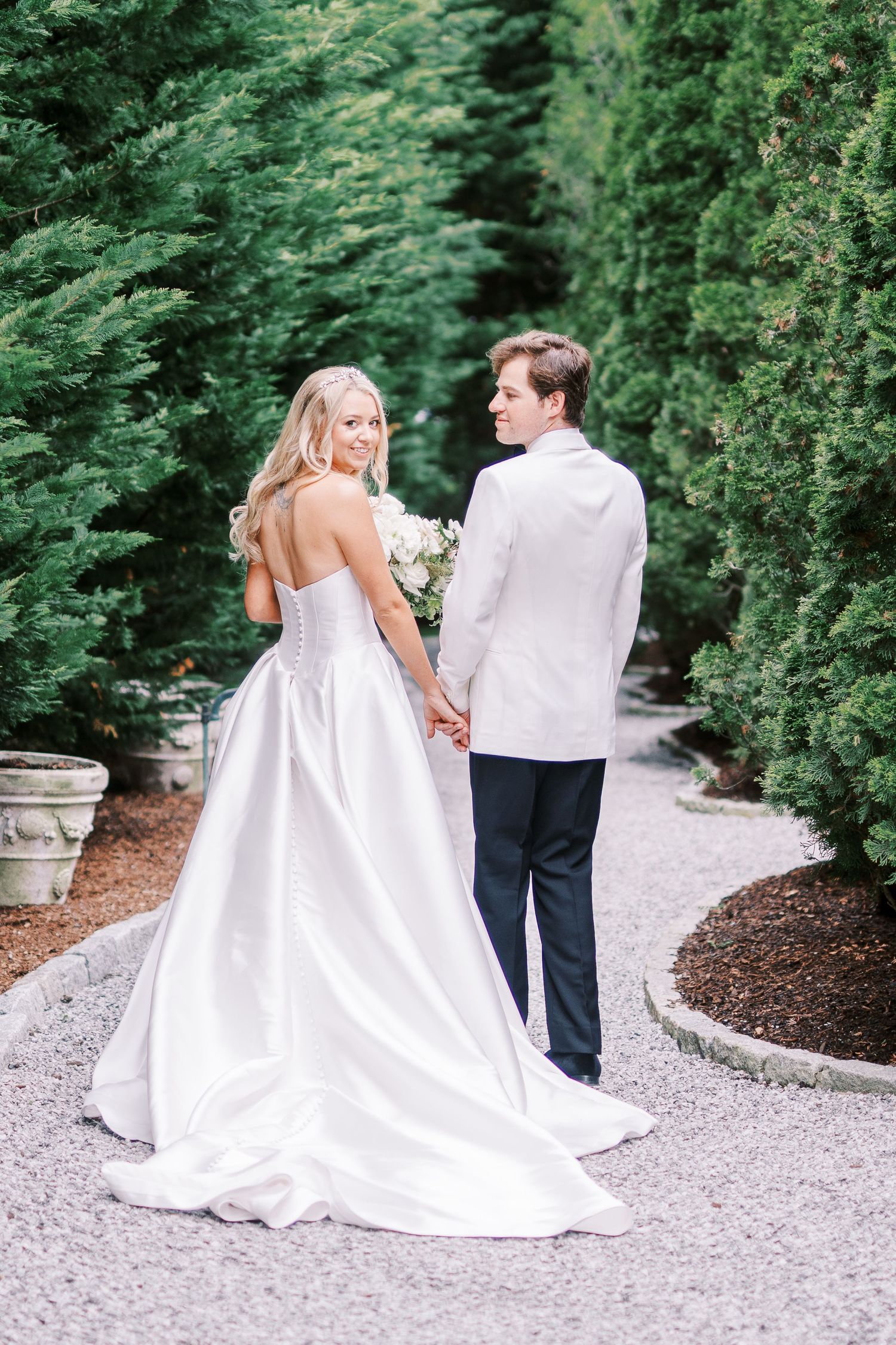 A couple in white wedding attire hold hands while walking along a gravel path surrounded by lush evergreen trees.