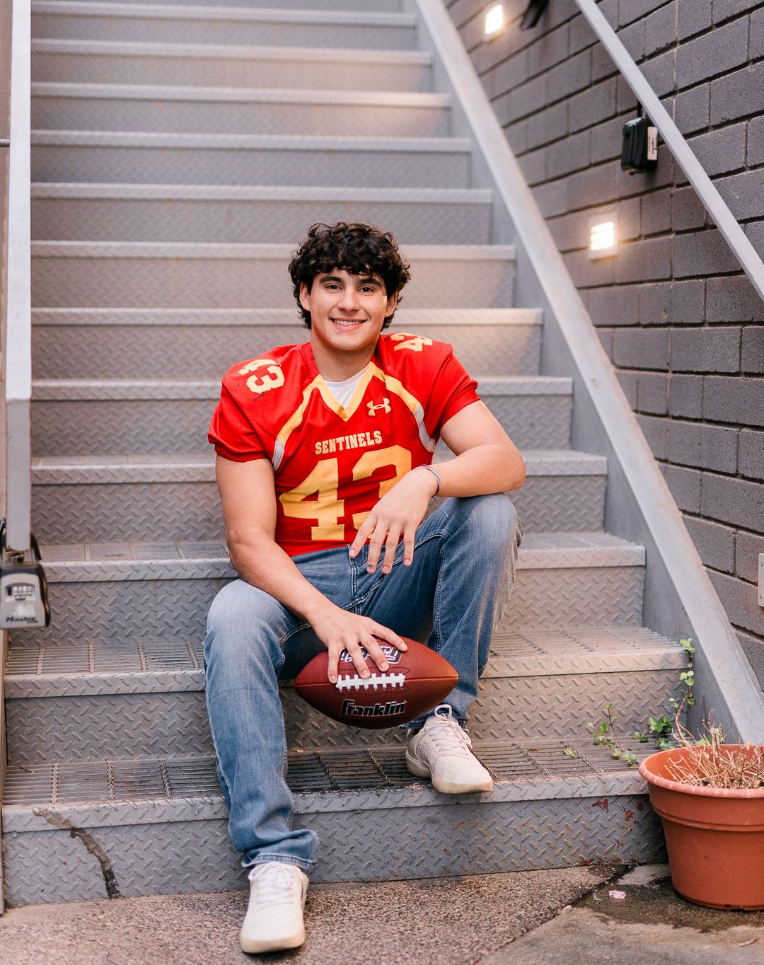 Student athlete in red football jersey number 43 sitting on outdoor stairs with football.