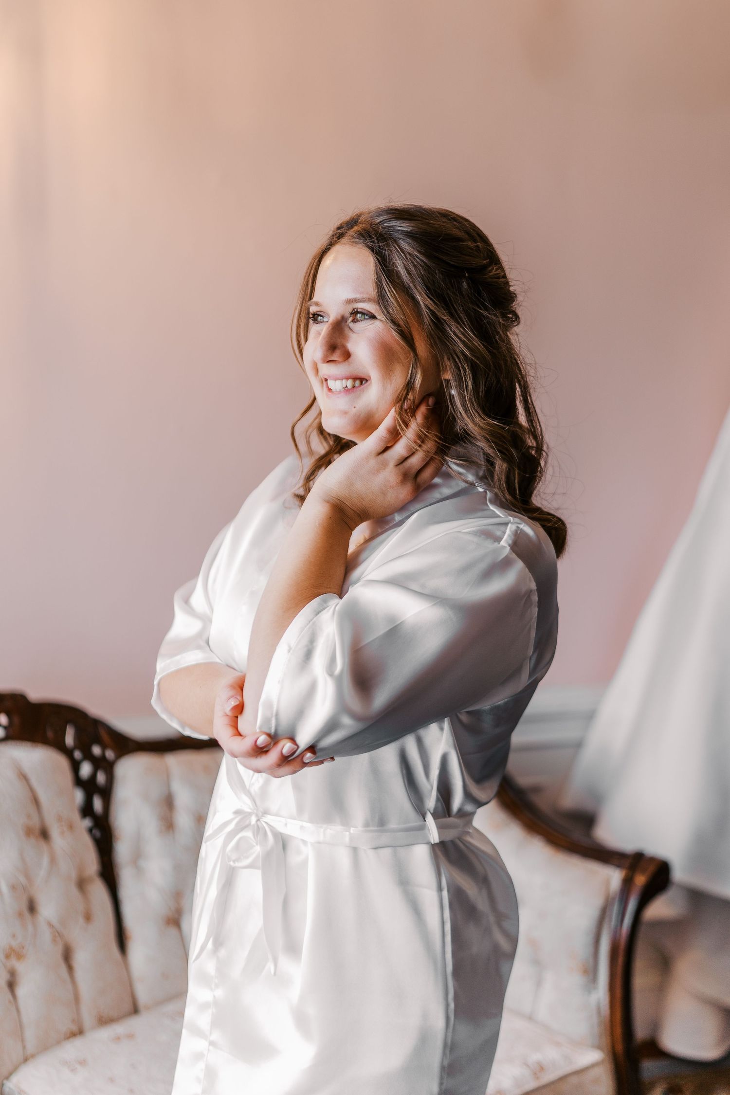 Someone in a white silk robe smiling and posing during a morning getting ready session against a pink wall.