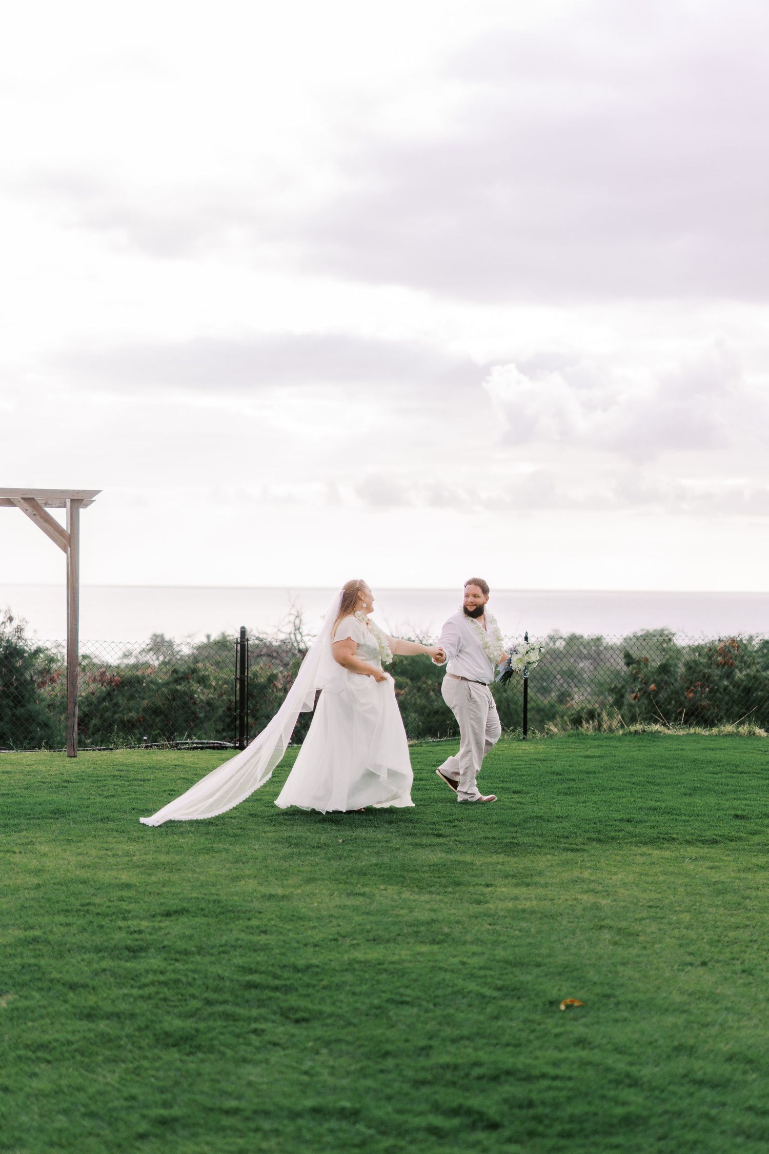 Newlyweds walking together on a grassy hilltop with wooden arch and cloudy sky in background.