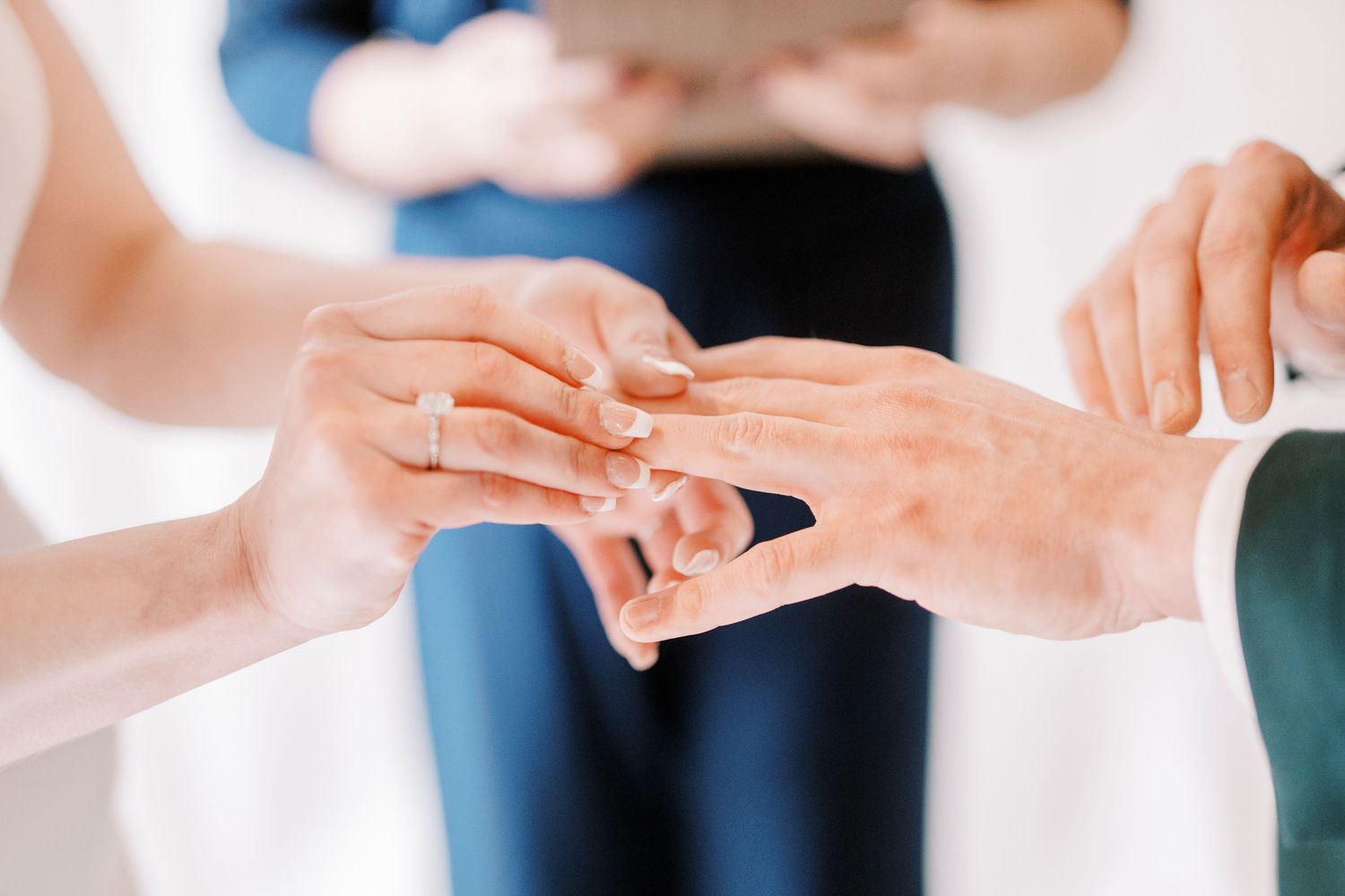 Close up of hands exchanging rings during a wedding ceremony against a soft white background.
