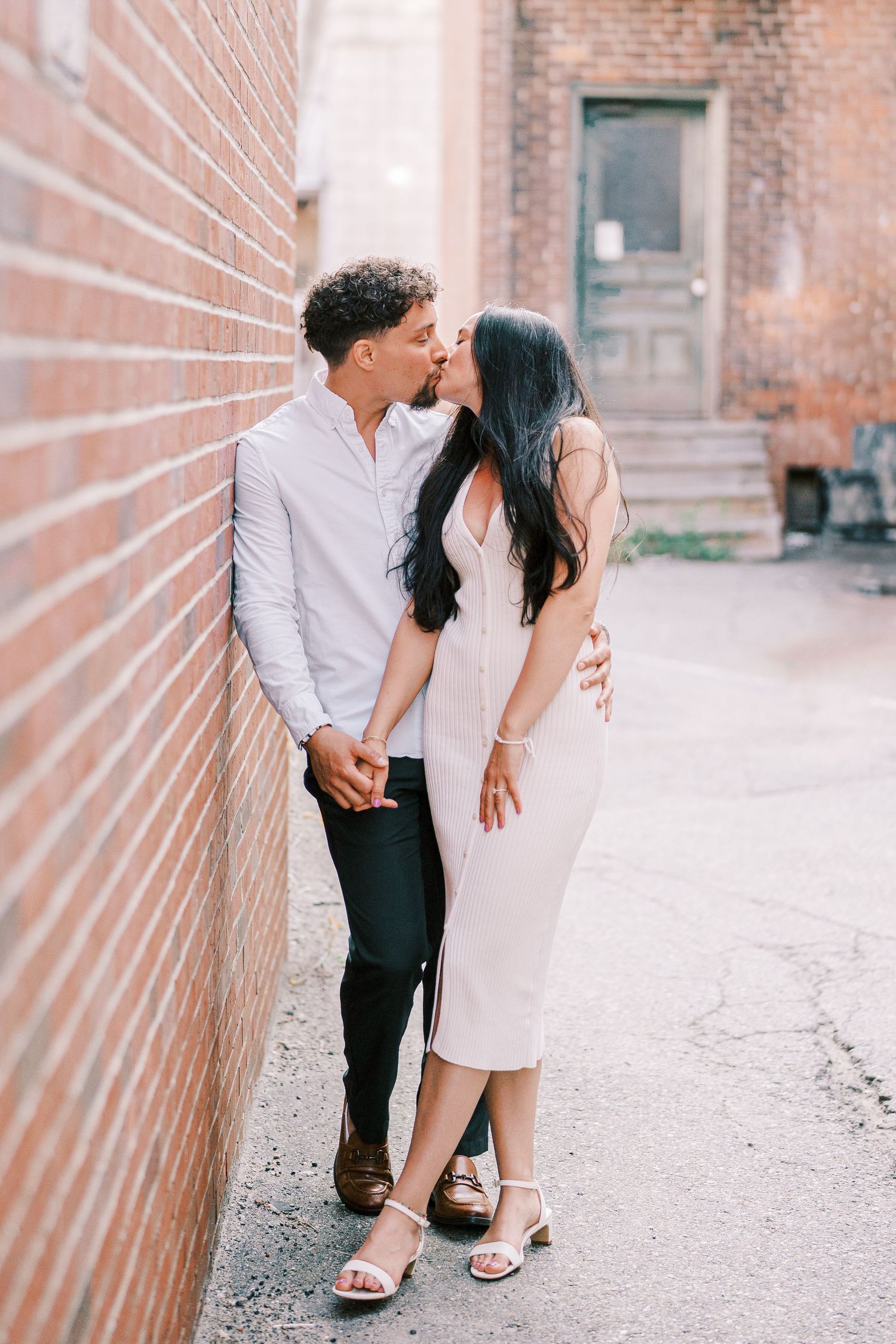 A couple in elegant attire shares an intimate moment against a brick wall backdrop.