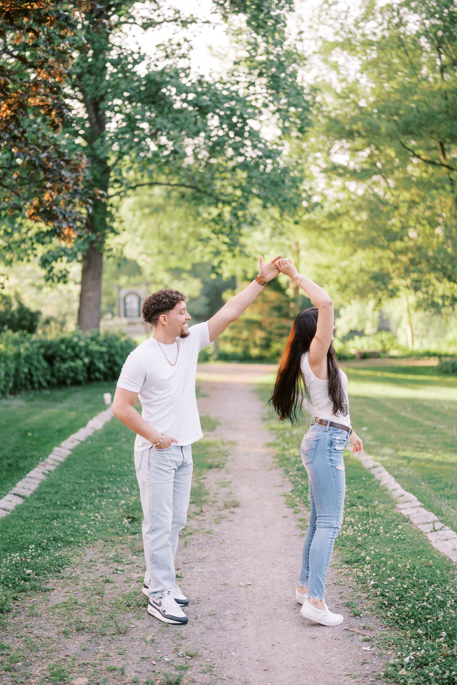 Two people in casual outfits dance playfully together on a tree-lined path in a sunny park setting.