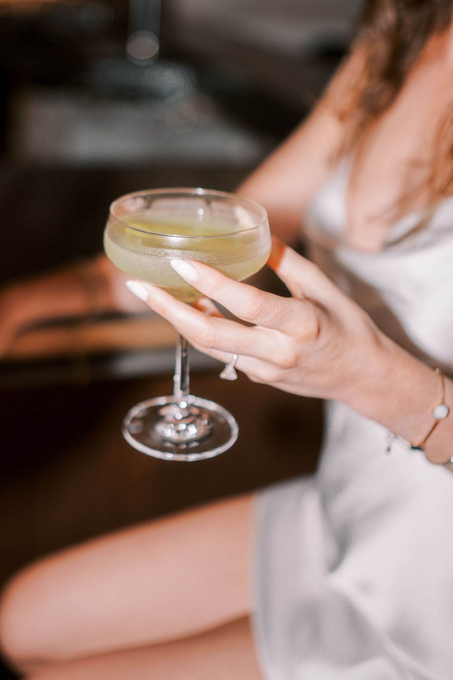 A hand with painted nails holds an elegant coupe glass filled with a pale yellow cocktail against a blurred background.