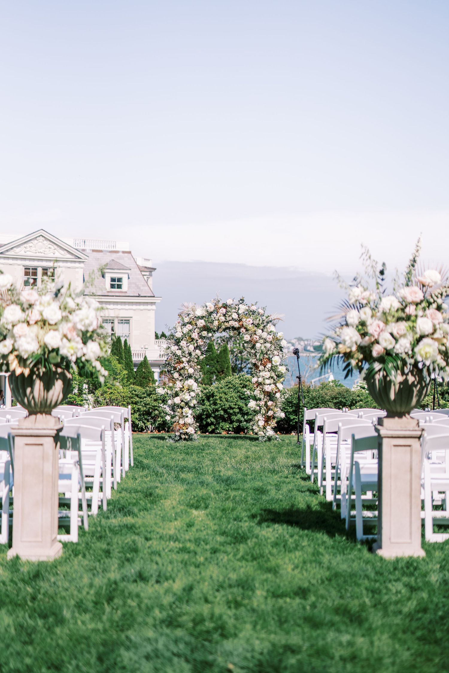 Elegant wedding ceremony setup with white chairs and pink floral arrangements along a grassy aisle overlooking a coastal view.