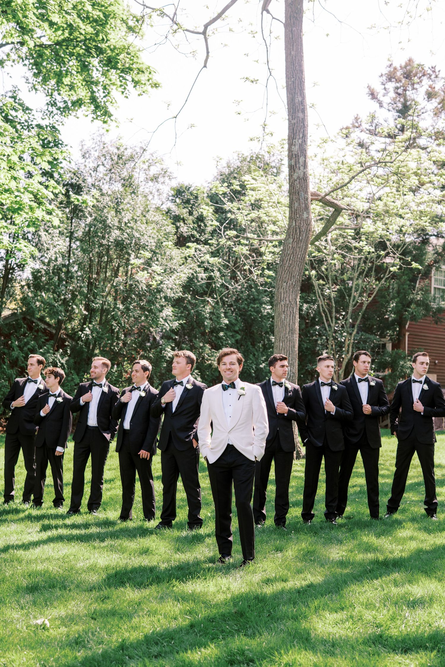 A long line of groomsmen in black tuxedos pose playfully on green lawn with trees in the background.