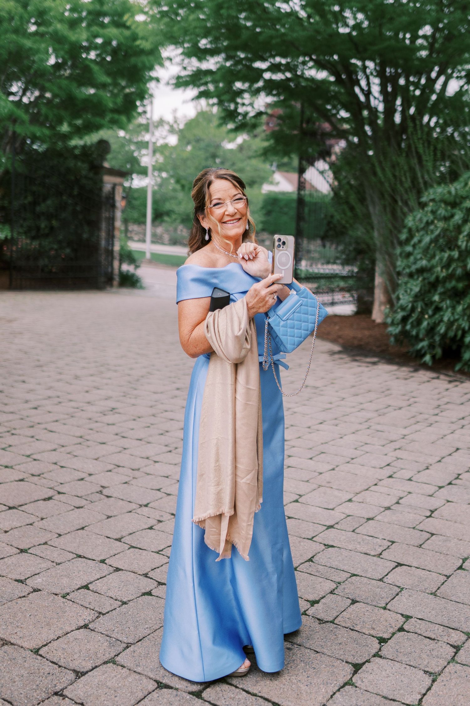 Woman in flowing blue off-shoulder gown poses on brick pathway surrounded by lush greenery.