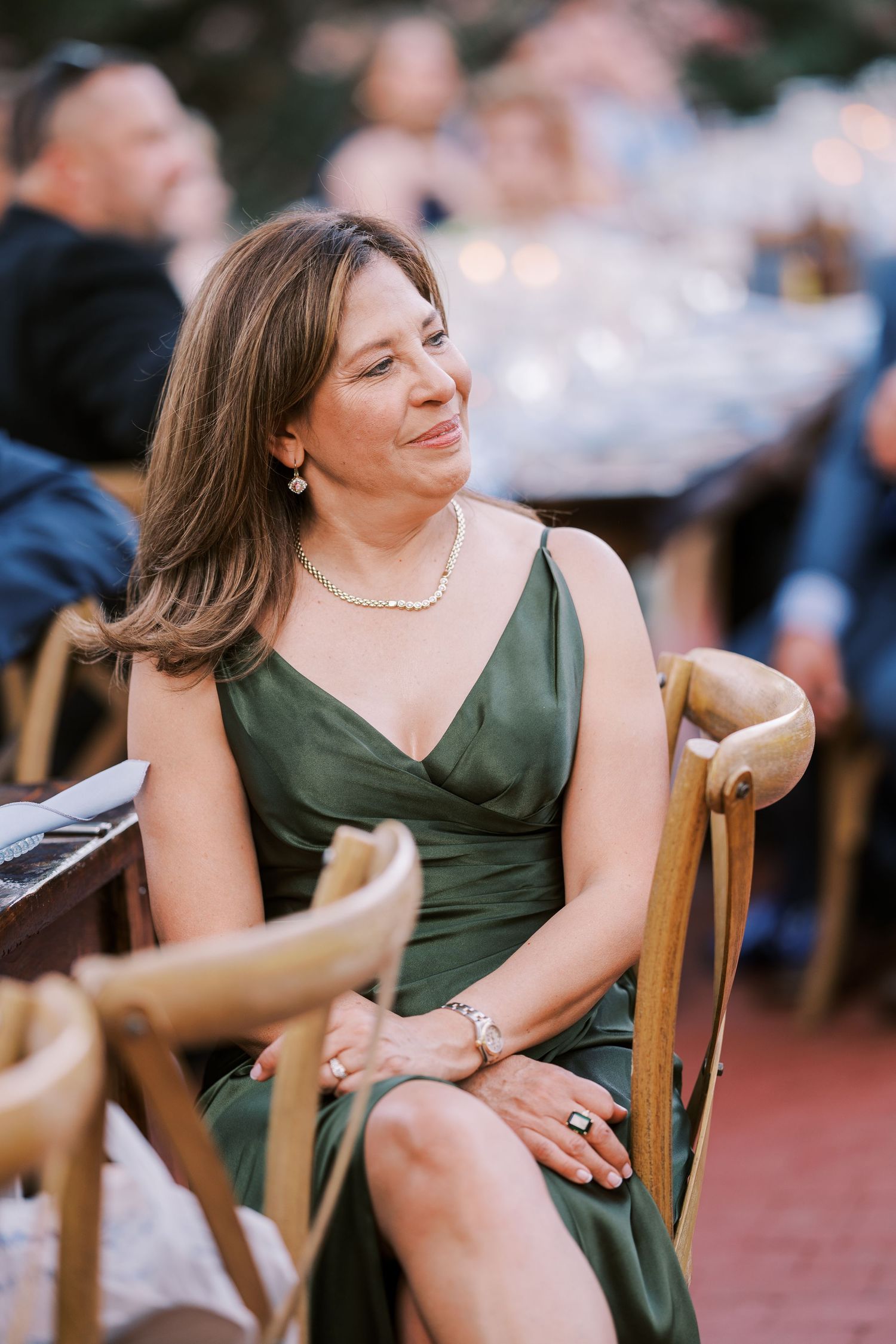 A guest in an elegant olive green dress and pearl necklace sits at an outdoor event.
