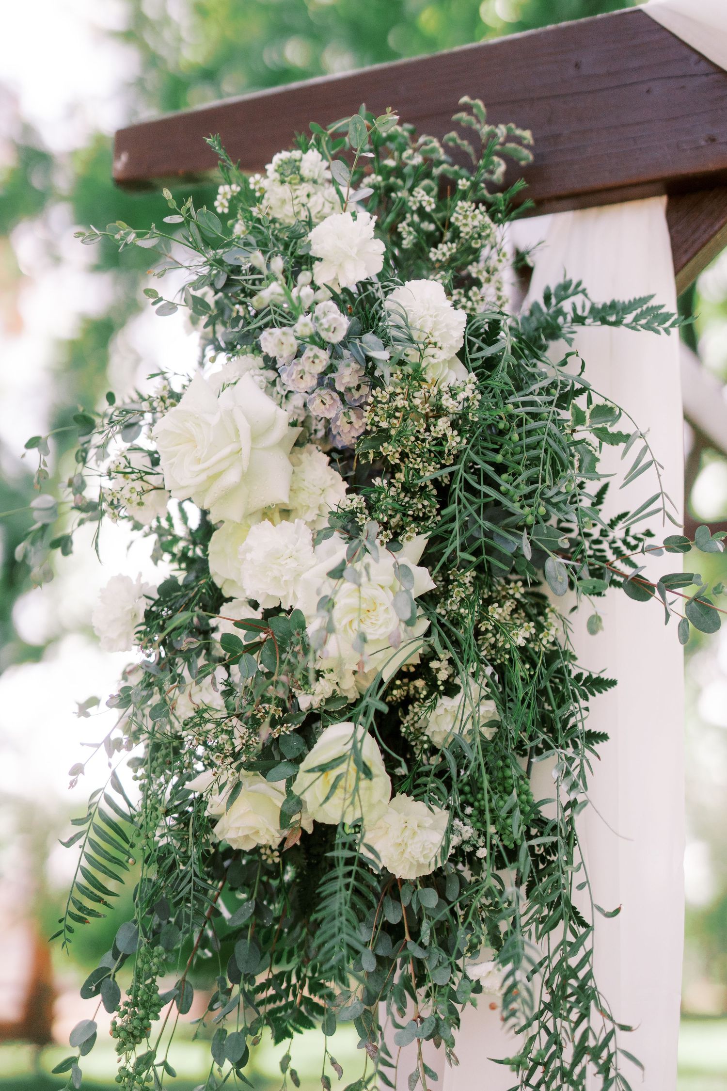 White floral arrangements with cascading greenery adorn a wooden pergola for an elegant outdoor wedding ceremony.