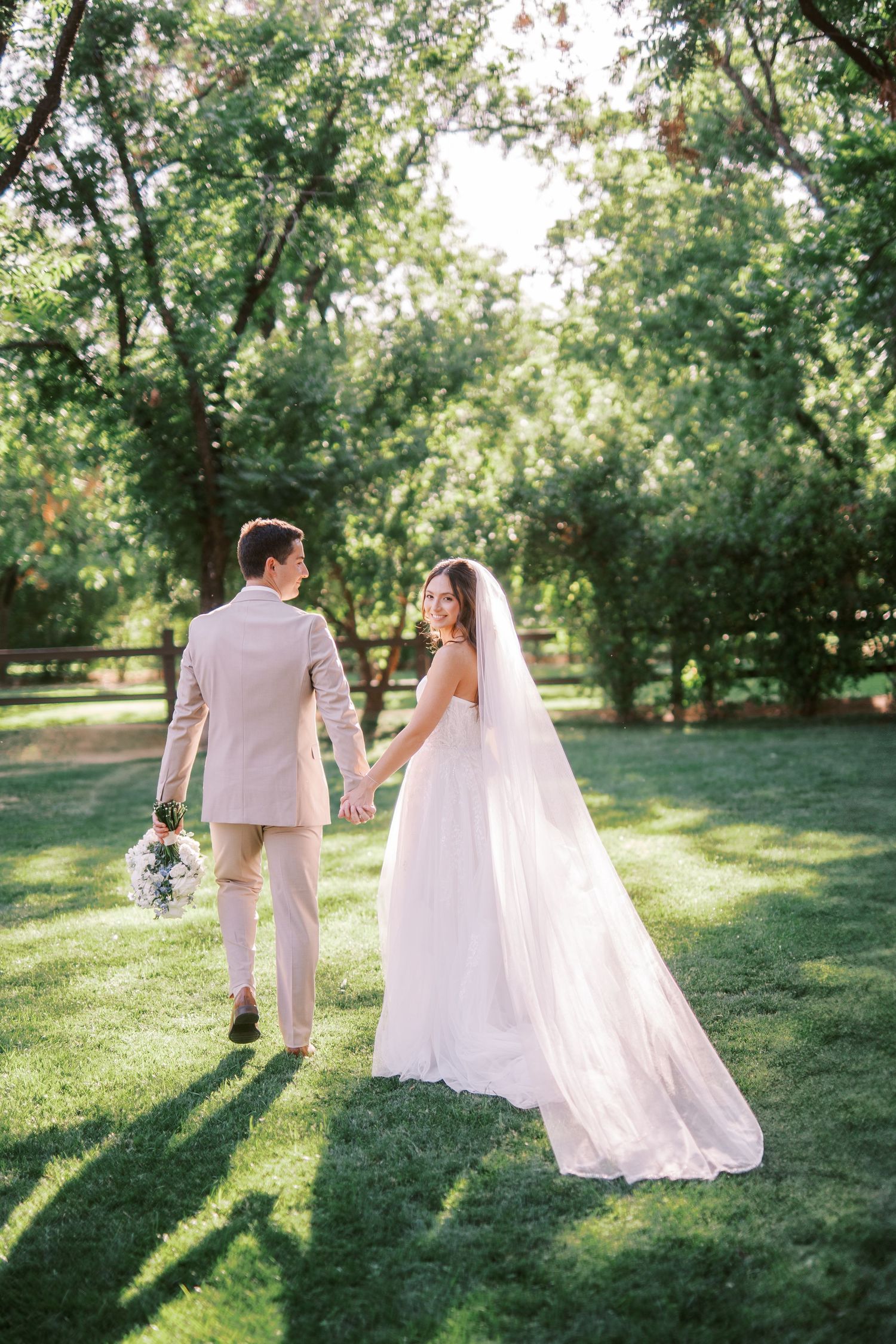 Newlywed couple walks hand in hand through a sun-dappled garden in warm afternoon light.