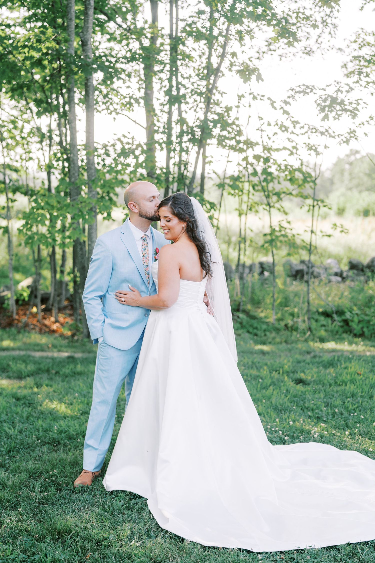 A couple in wedding attire embrace in a romantic outdoor setting with trees and greenery in the background.