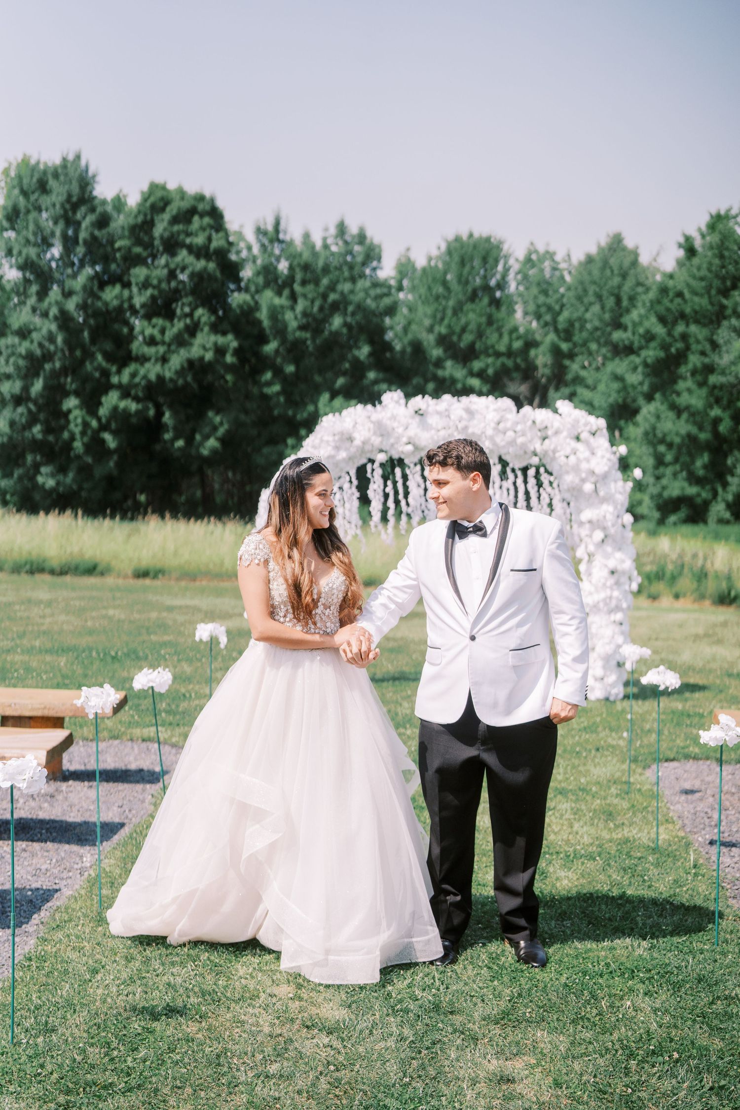 Wedding couple stands in front of white floral arch on sunny lawn with trees in background.