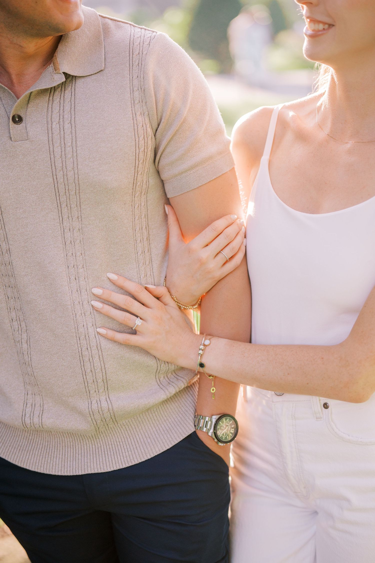 A couple wearing neutral tones embrace in soft natural lighting showing engagement rings and casual style.