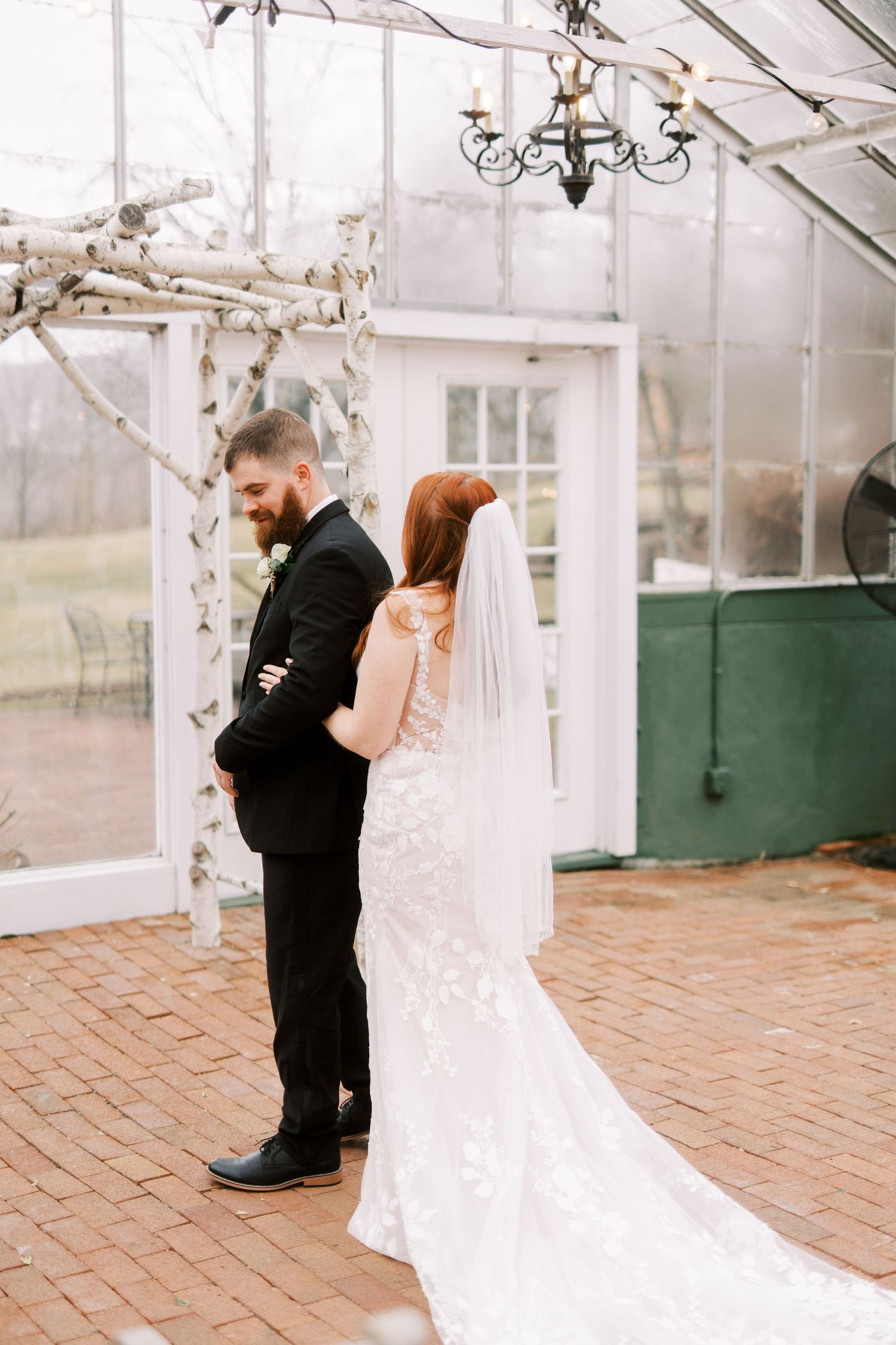 A couple shares an intimate first look moment in a sunlit greenhouse with brick floors and white decor.