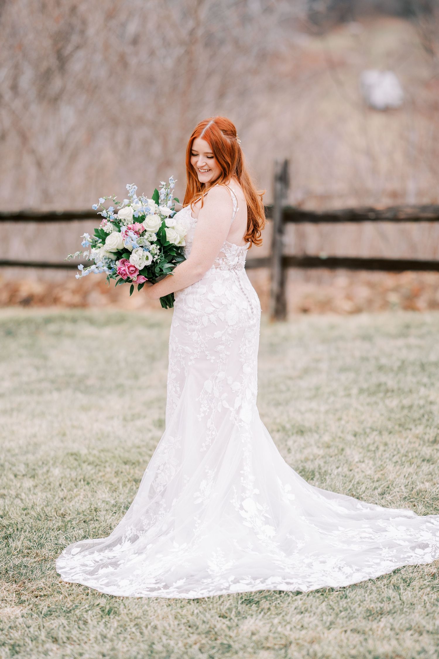 Bride in lace wedding dress with long train holding pink and white bouquet in outdoor garden setting.