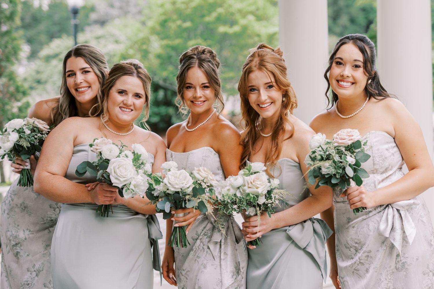 Bridesmaids in light gray strapless gowns hold white and green wedding bouquets while posing outdoors.