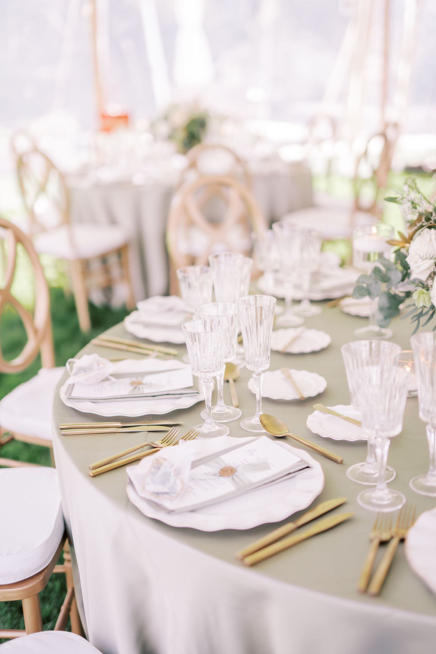 Table settings for an elegant wedding reception with gold cutlery, crystal glasses and white china on a neutral tablecloth.