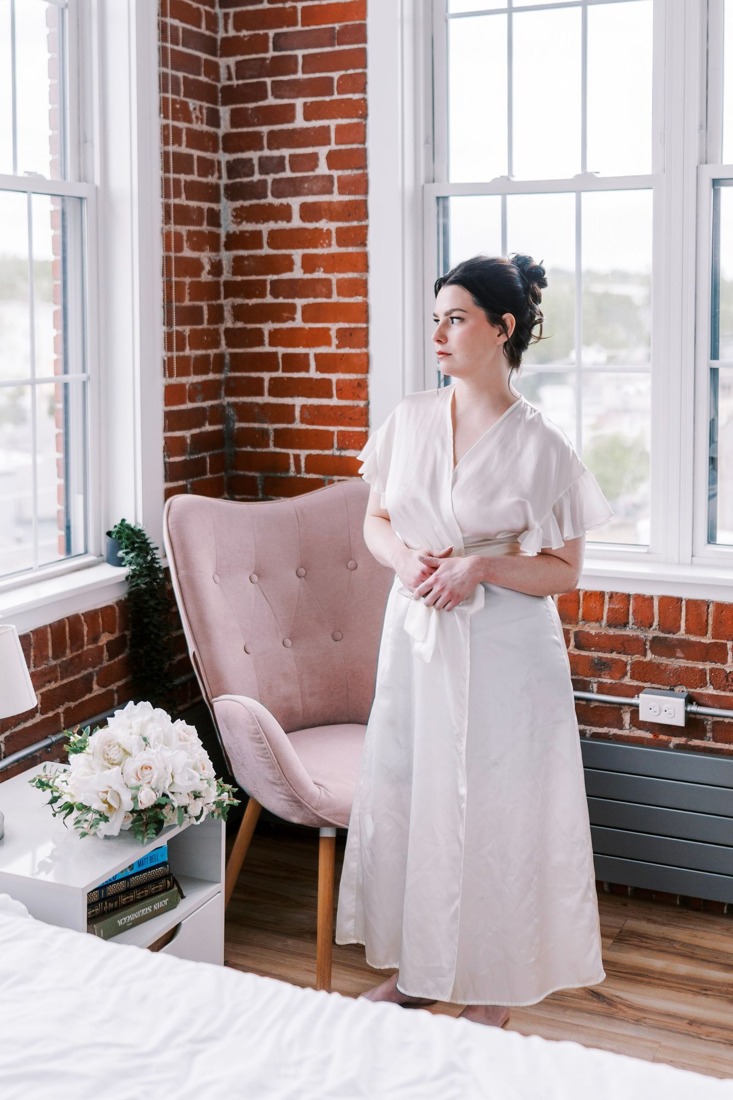 Woman in white robe stands by pink tufted chair near exposed brick wall and bright windows in elegant bedroom setting.